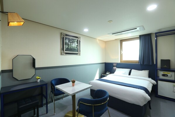 Quarto de hotel com cama, escrivaninha, duas cadeiras, uma mesa pequena com uma planta, um quadro na parede e uma janela com cortinas.