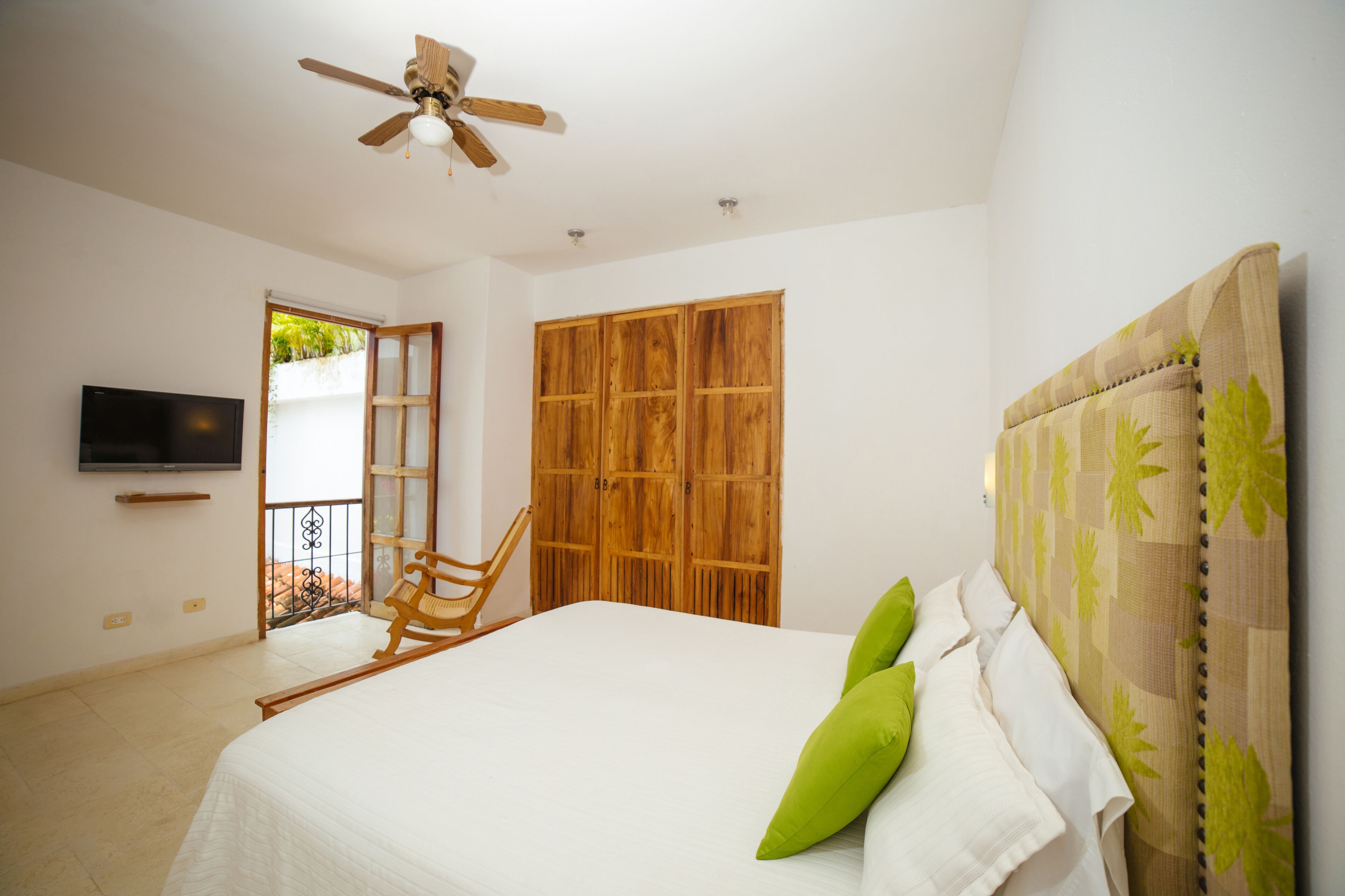 Un dormitorio con una cama, un armario de madera, un ventilador de techo y un televisor en la pared.