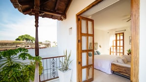 Un balcón con una planta en maceta, una cama con ropa de cama blanca y una puerta de madera que da a una habitación.