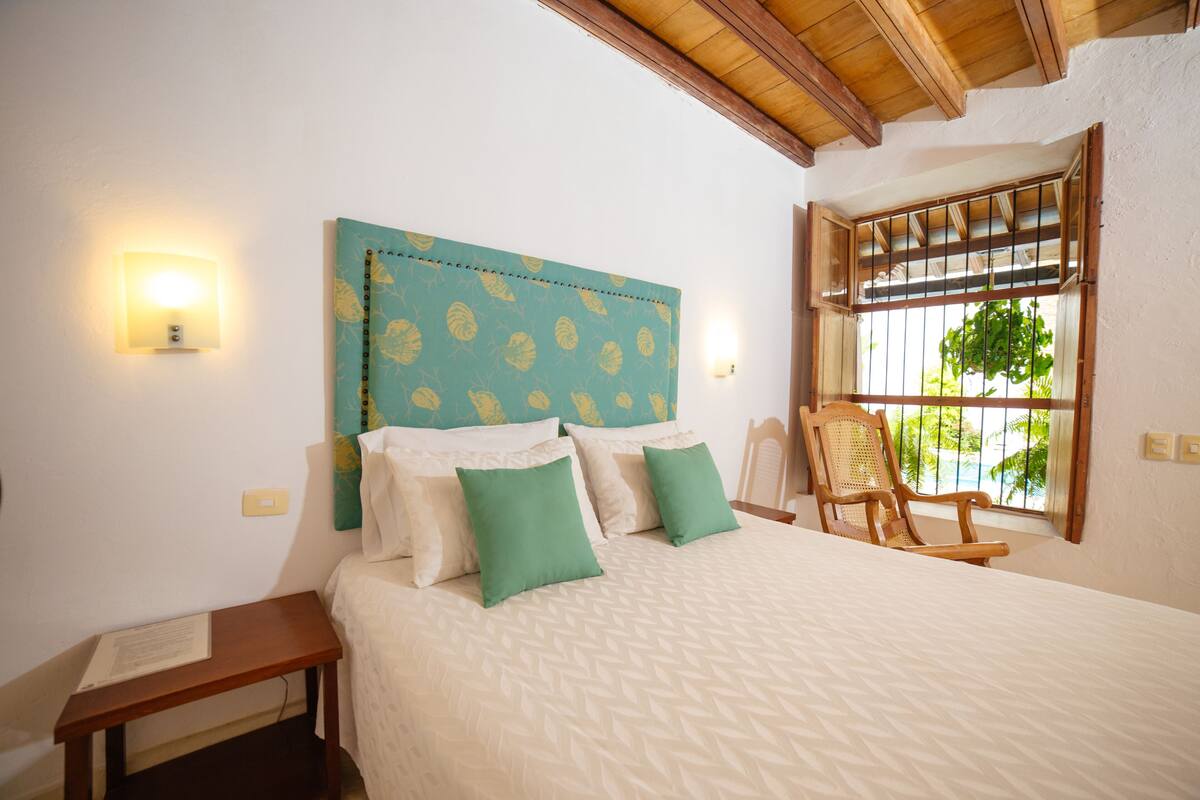 Een slaapkamer met een bed, kussens, een nachtkastje, een stoel, een raam met uitzicht op groen en een houten plafond.