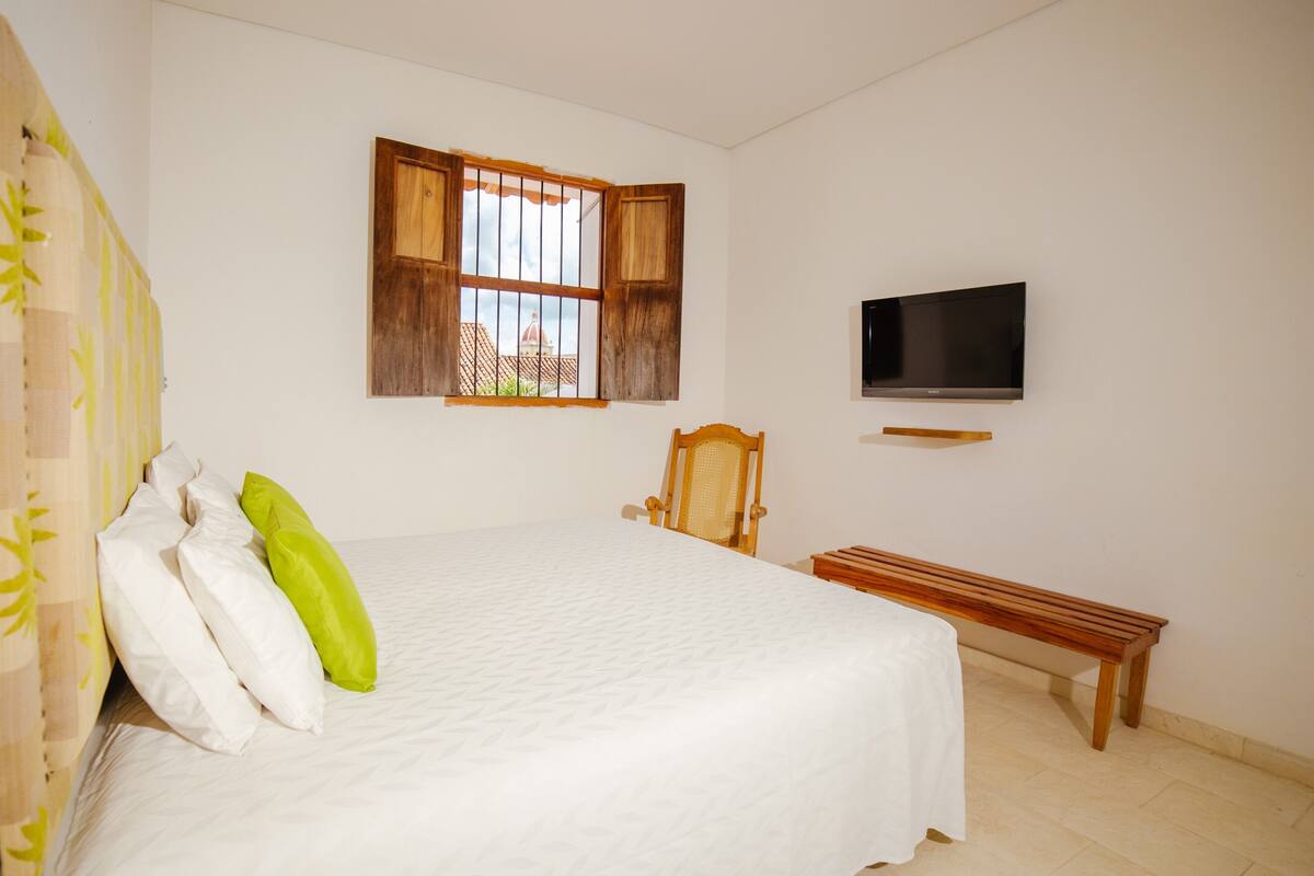 Een slaapkamer met een groot bed, een houten stoel, een bankje, een aan de muur bevestigde televisie en een raam met houten luiken.