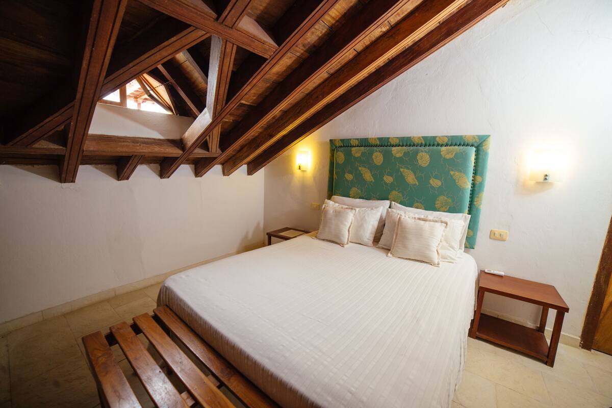 Een slaapkamer met een houten plafond, een bed met een groen hoofdeinde, twee nachtkastjes en een bankje aan het voeteneind van het bed.