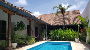 Una piscina con bordes de azulejos, rodeada de plantas en macetas y una pérgola con una hamaca.
