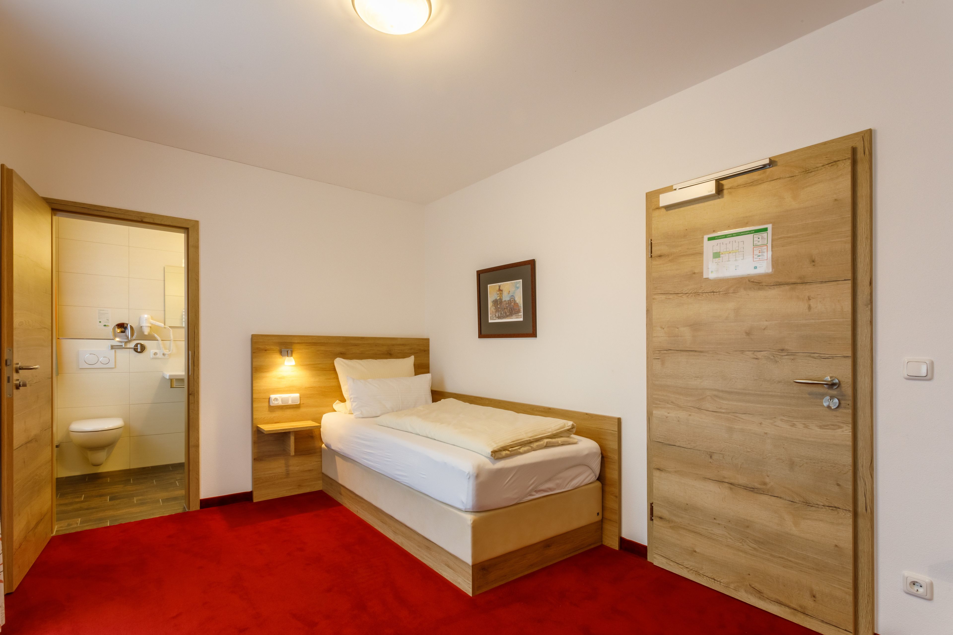 Habitación de hotel con una cama de madera, una mesita de noche con lámpara, una puerta de madera y una alfombra roja.