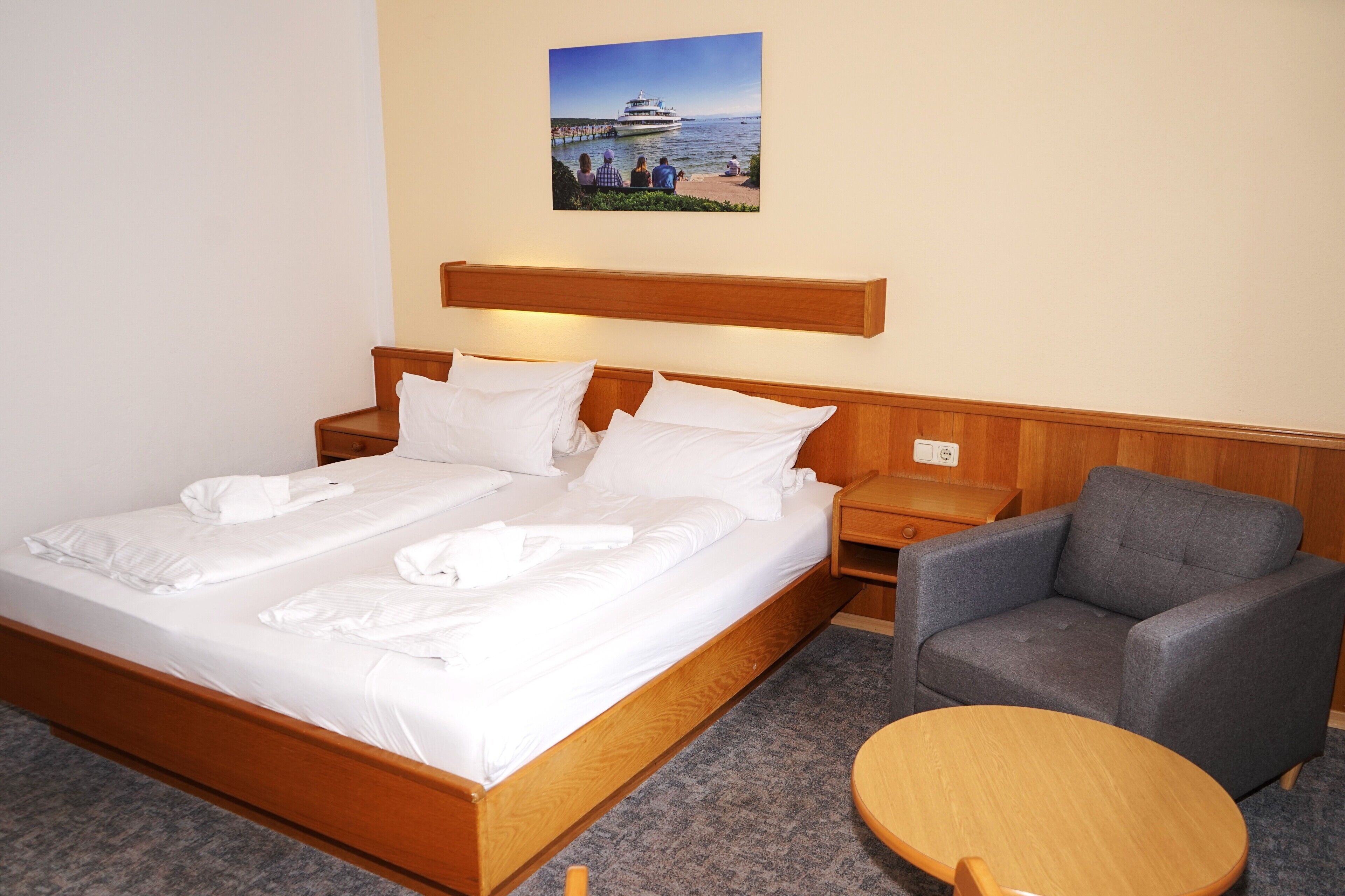 Habitación de hotel con una cama de madera, un sillón gris, una mesa redonda de madera y un cuadro de un crucero en la pared.