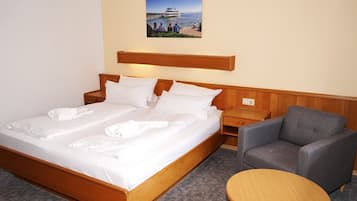 Habitación de hotel con una cama de madera, un sillón gris, una mesa redonda de madera y un cuadro de un crucero en la pared.