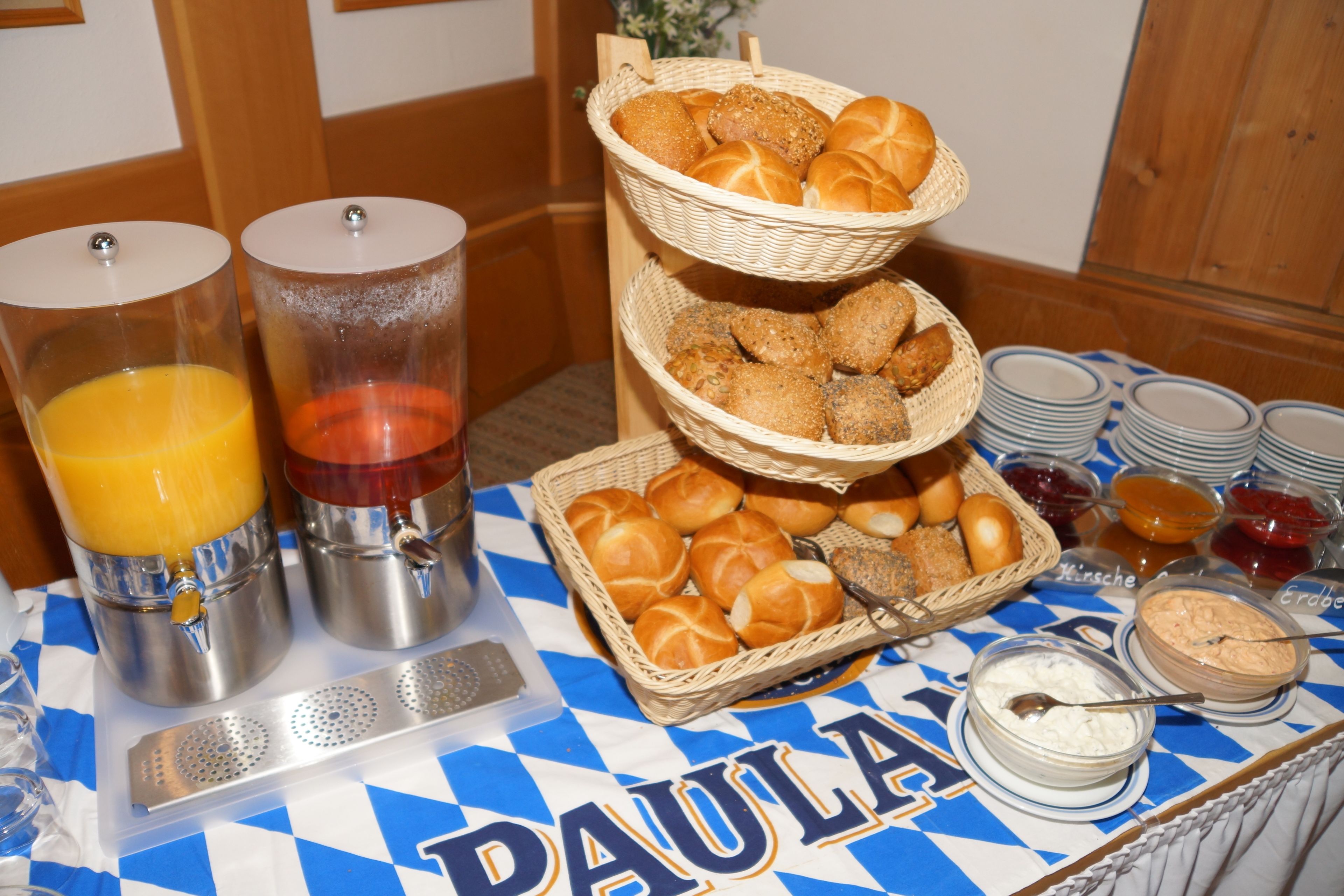 Una mesa de buffet con mantel azul y blanco a cuadros, dos dispensadores de jugo, cestas de pan y diferentes condimentos.
