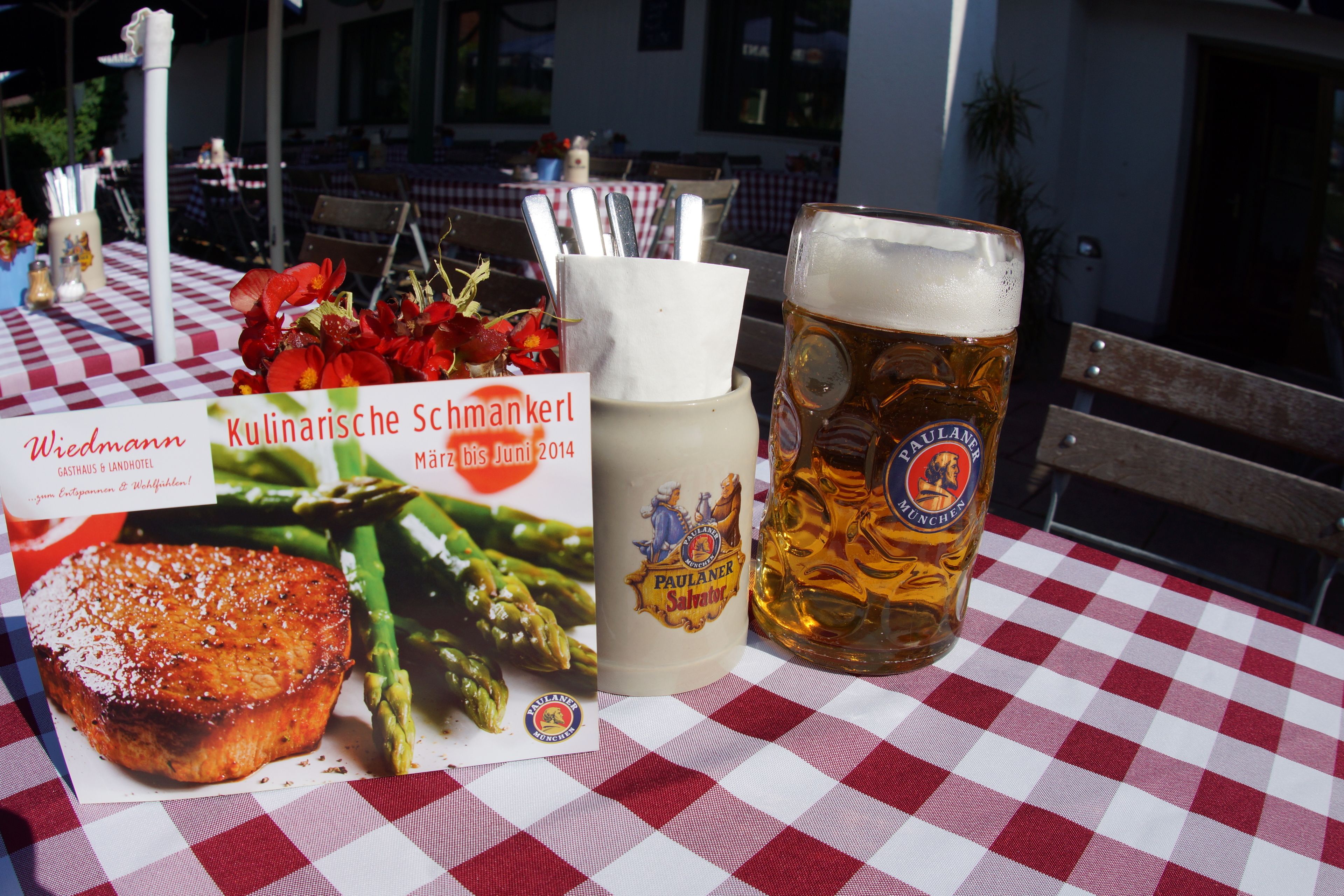 A table with a red and white checkered tablecloth, a beer glass, a bottle of beer, and a menu with asparagus and meat.