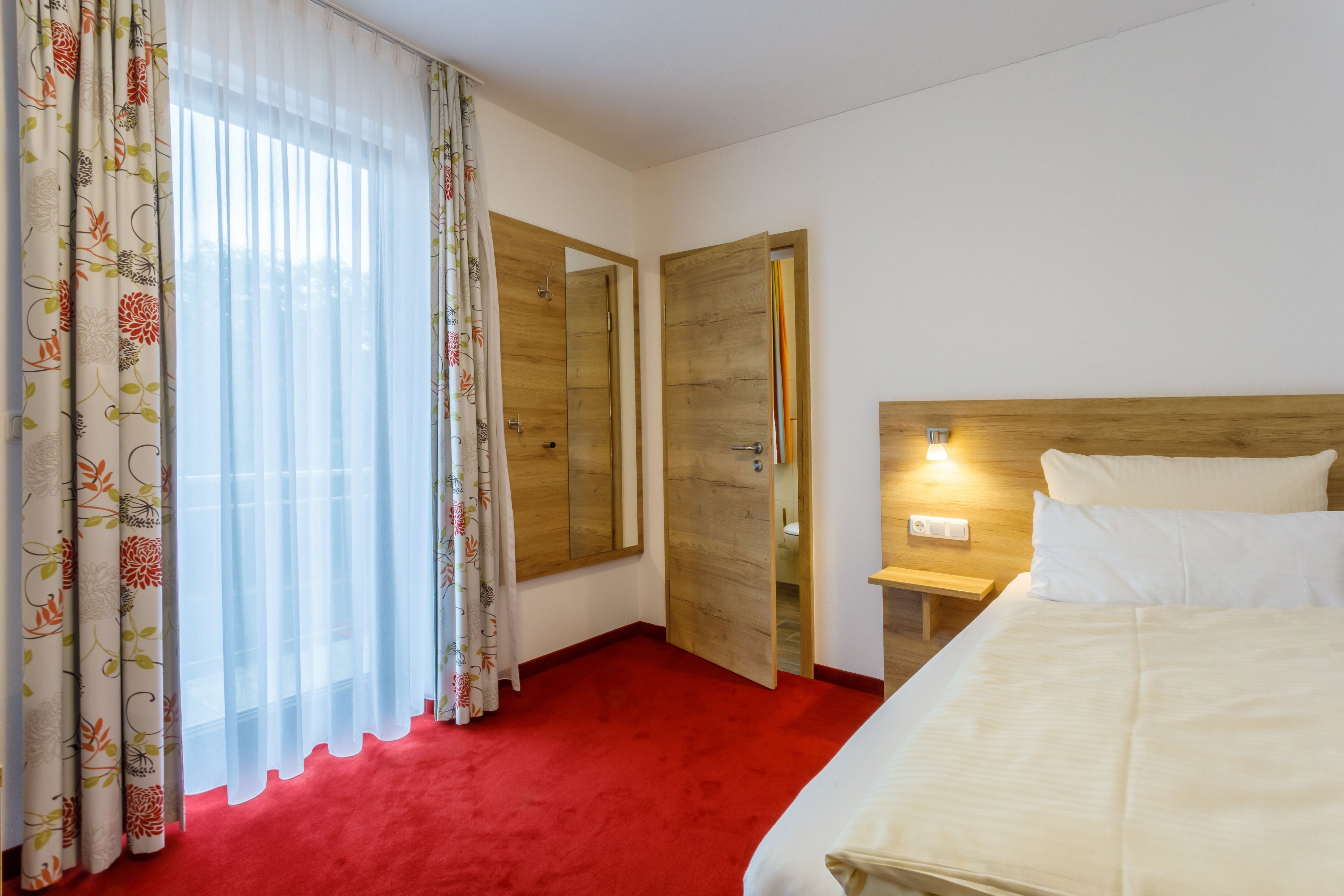 A hotel room with a red carpet, a bed with white linens, a wooden headboard, a nightstand with a lamp, and a large mirror on the wall.
