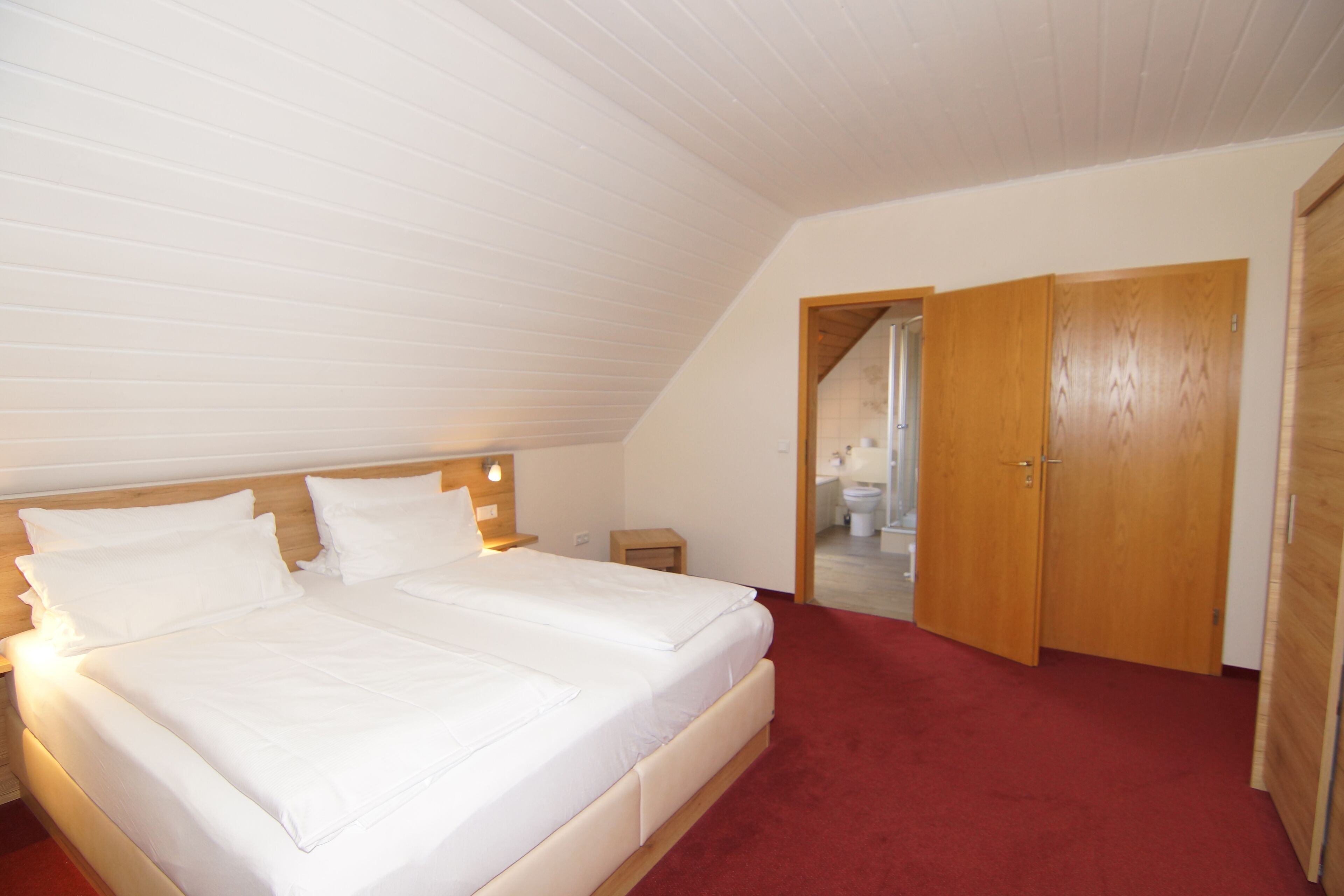 Un dormitorio con una cama grande, cabecera de madera y alfombra roja. Se observa una puerta de madera y una mesita de noche con lámpara.