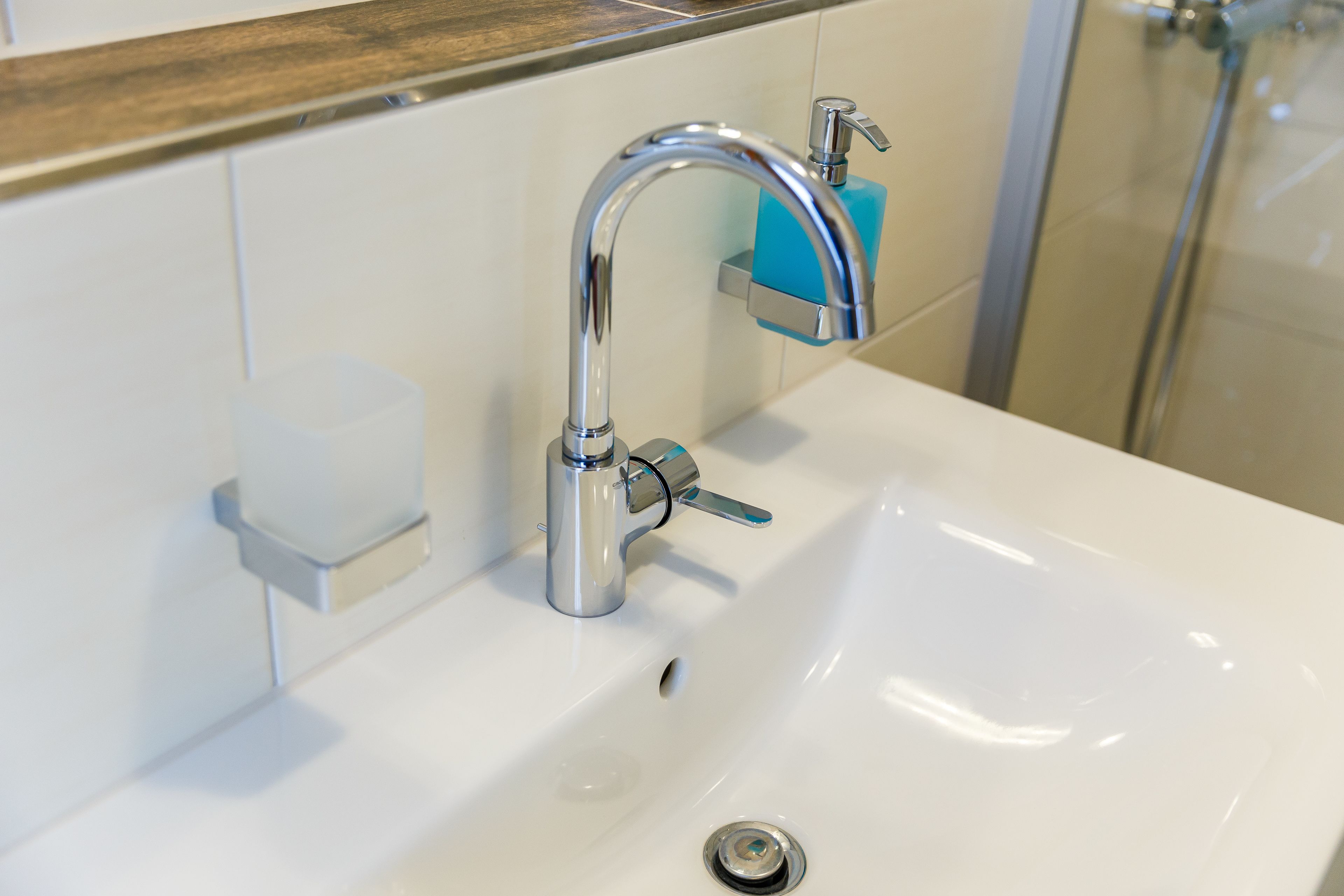 A modern bathroom with a white sink, chrome faucet, and a soap dispenser on the wall.