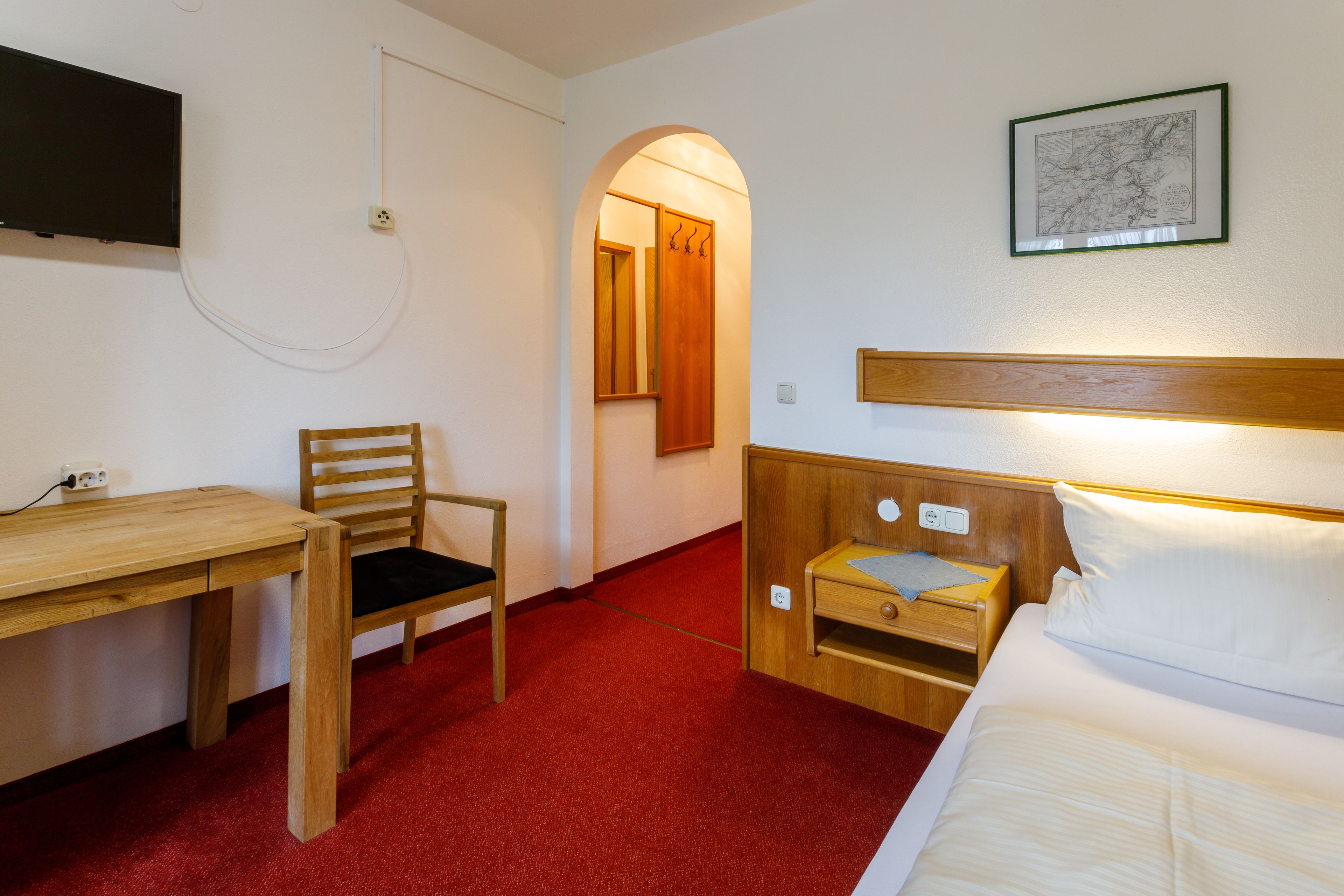 A hotel room with a wooden desk, a chair, a bed with a wooden headboard, a nightstand, and a framed picture on the wall.