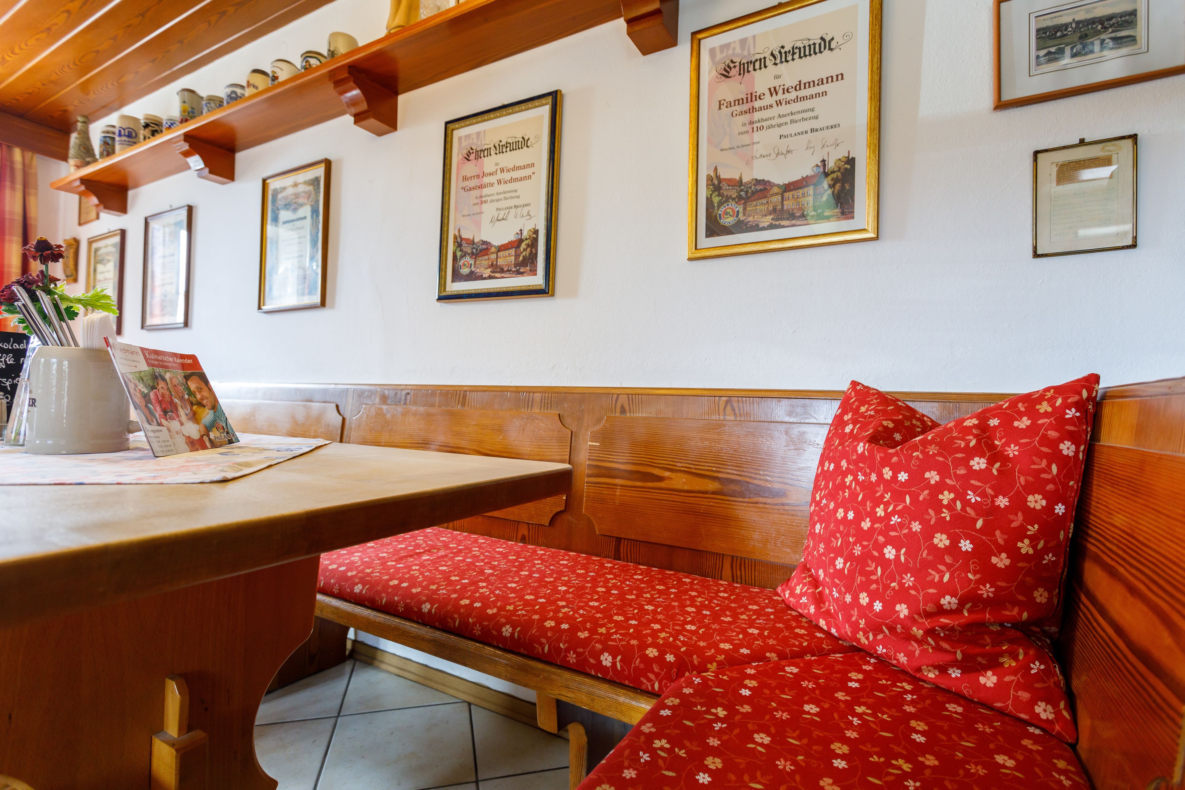 Eine hölzerne Bank mit rotem Blumenkissen, ein Holztisch mit weißer Karaffe und einer Speisekarte, sowie gerahmte Zertifikate an der Wand.