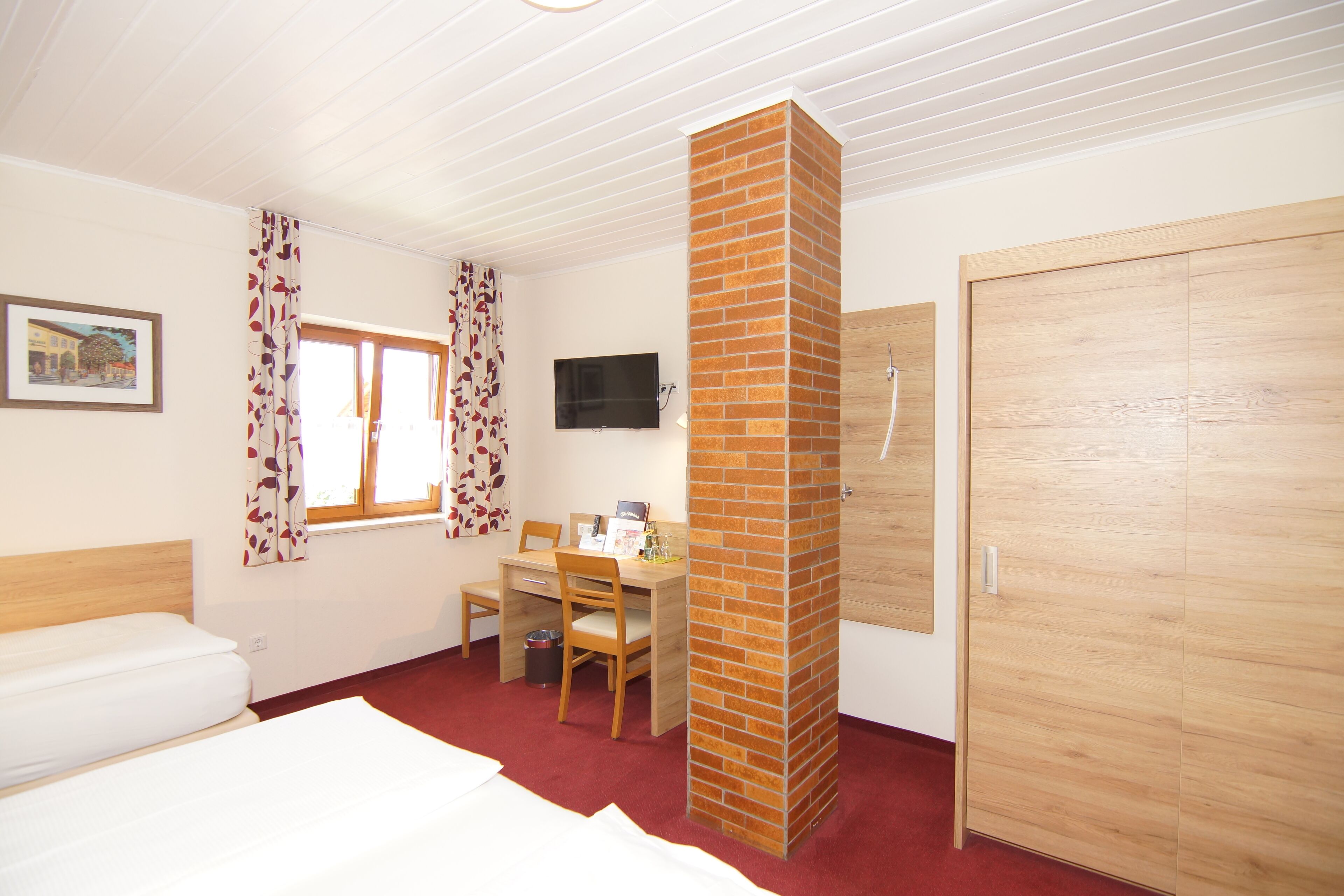 A bedroom with a bed, a desk, a chair, a TV, a wooden wardrobe, and a brick fireplace.