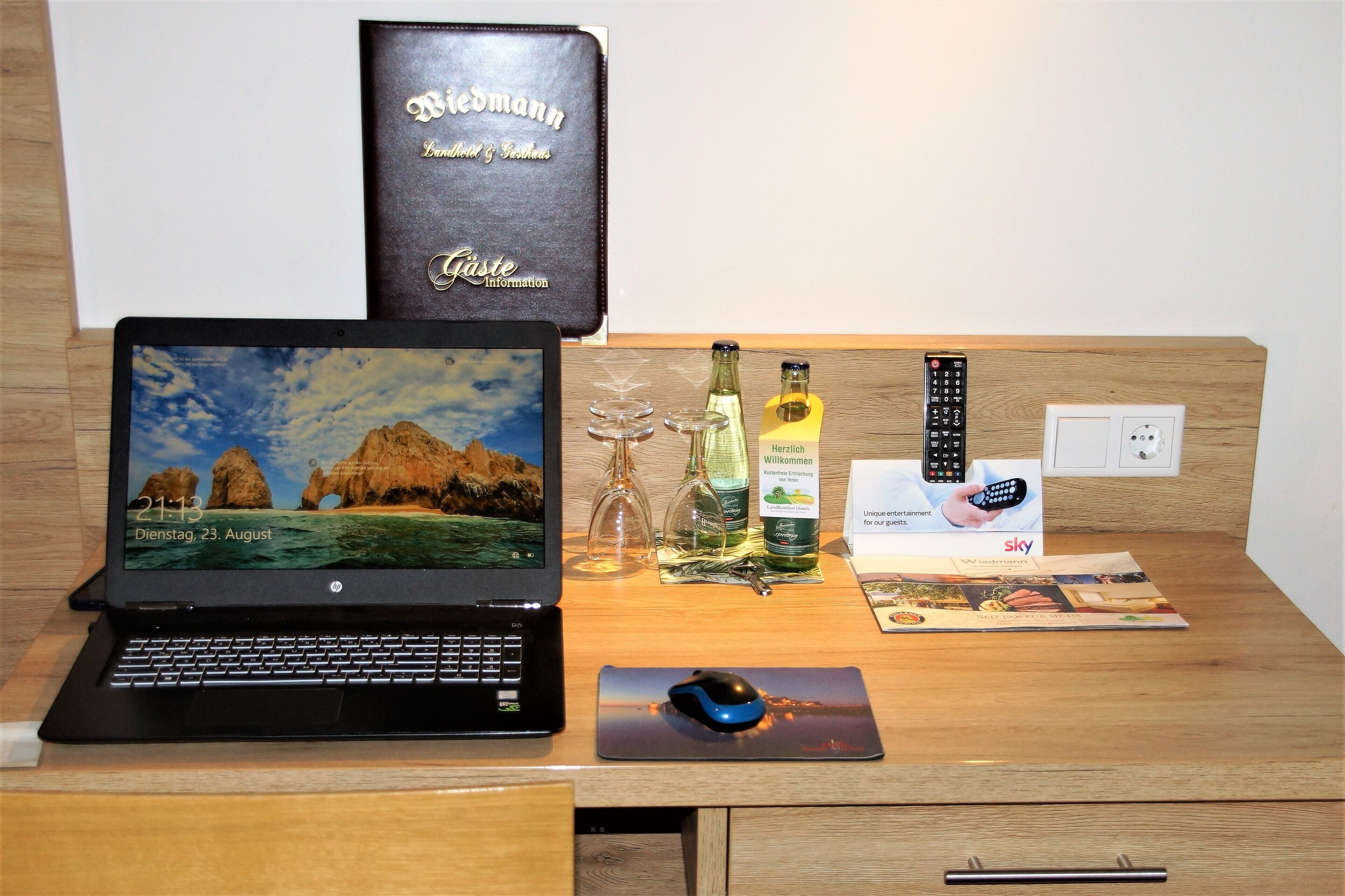 Una laptop sobre un escritorio de madera con un fondo de pantalla de paisaje, una botella de vino, un control remoto y una revista Sky.