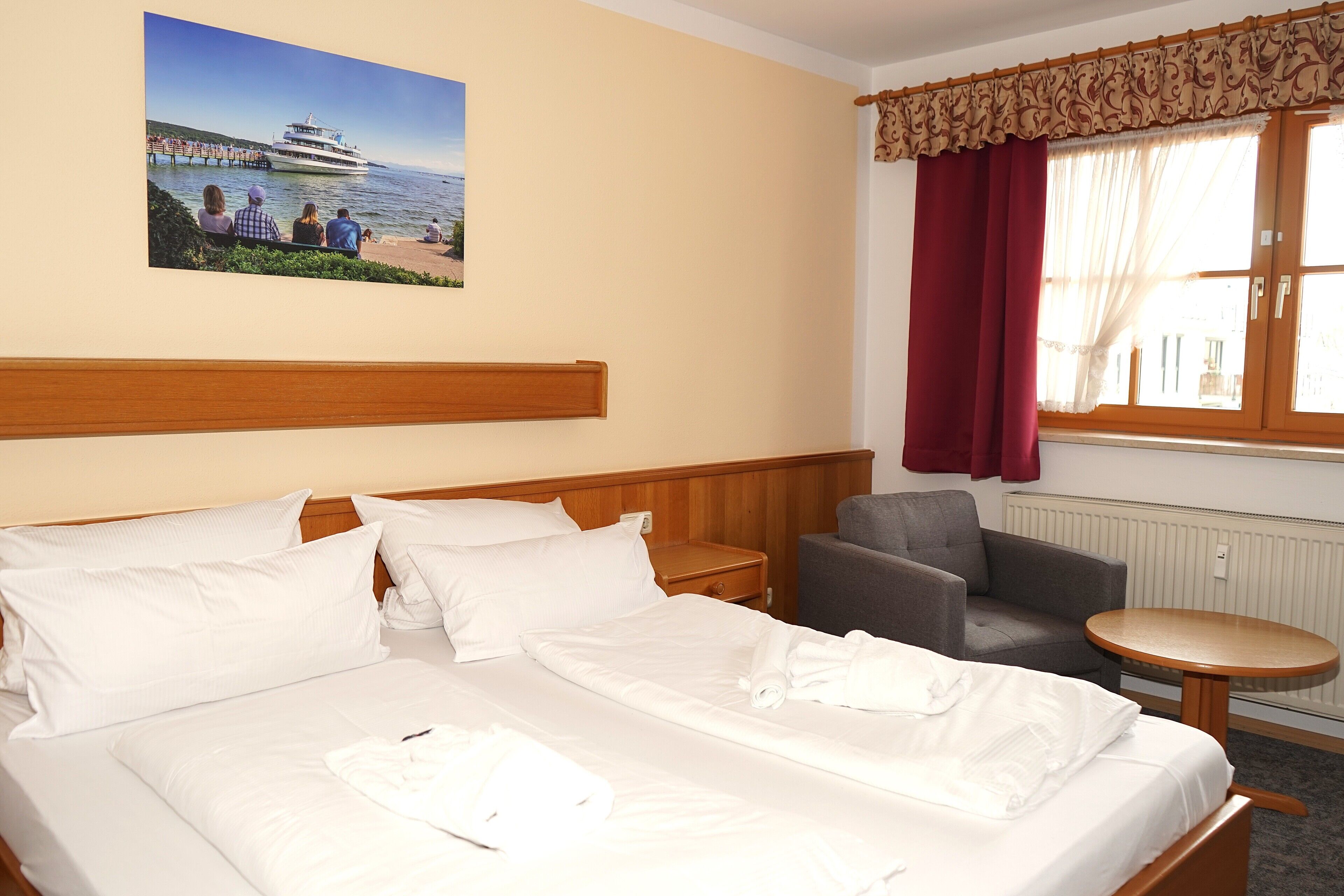 A hotel room with a bed, a chair, a small table, a nightstand, a window with curtains, and a painting of a pier.