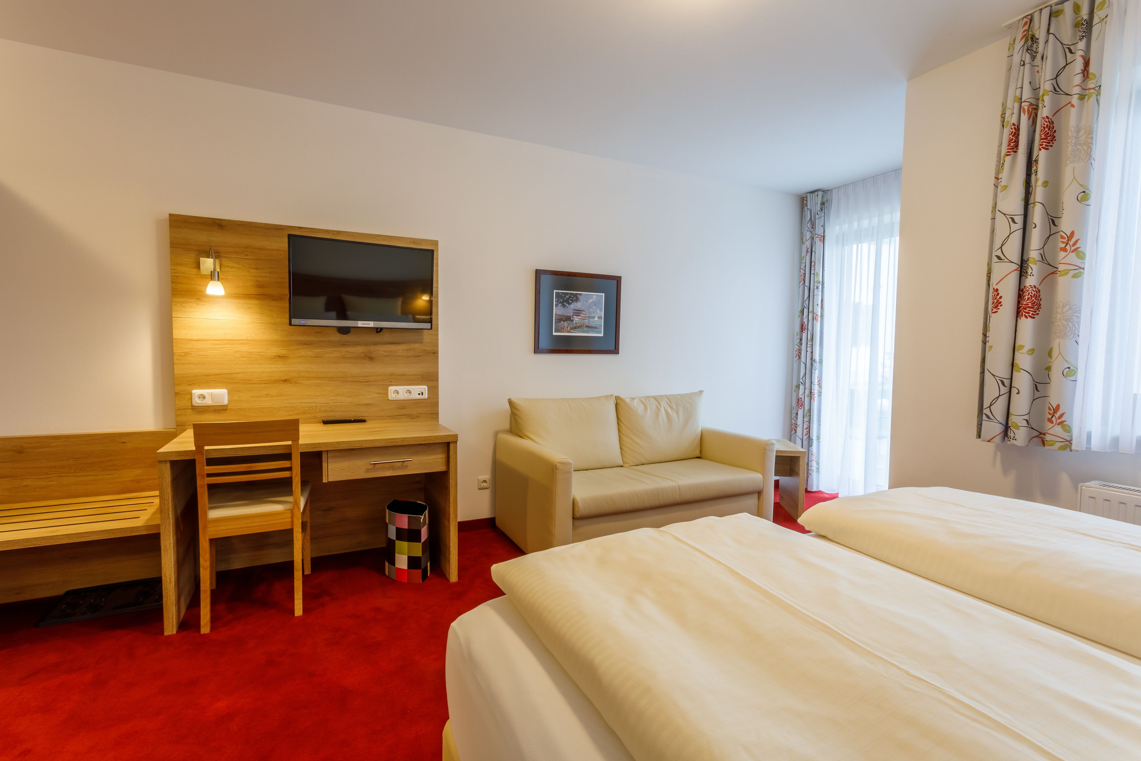 Habitación de hotel con cama, escritorio, silla, sofá, televisor montado en una pared de madera y ventana con cortinas.