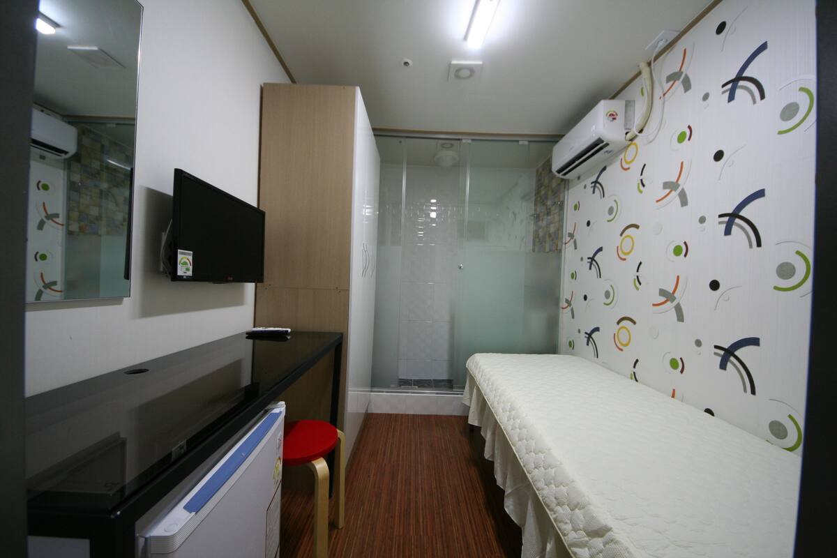A small, compact room with a bed, a desk, a chair, and a shower area.