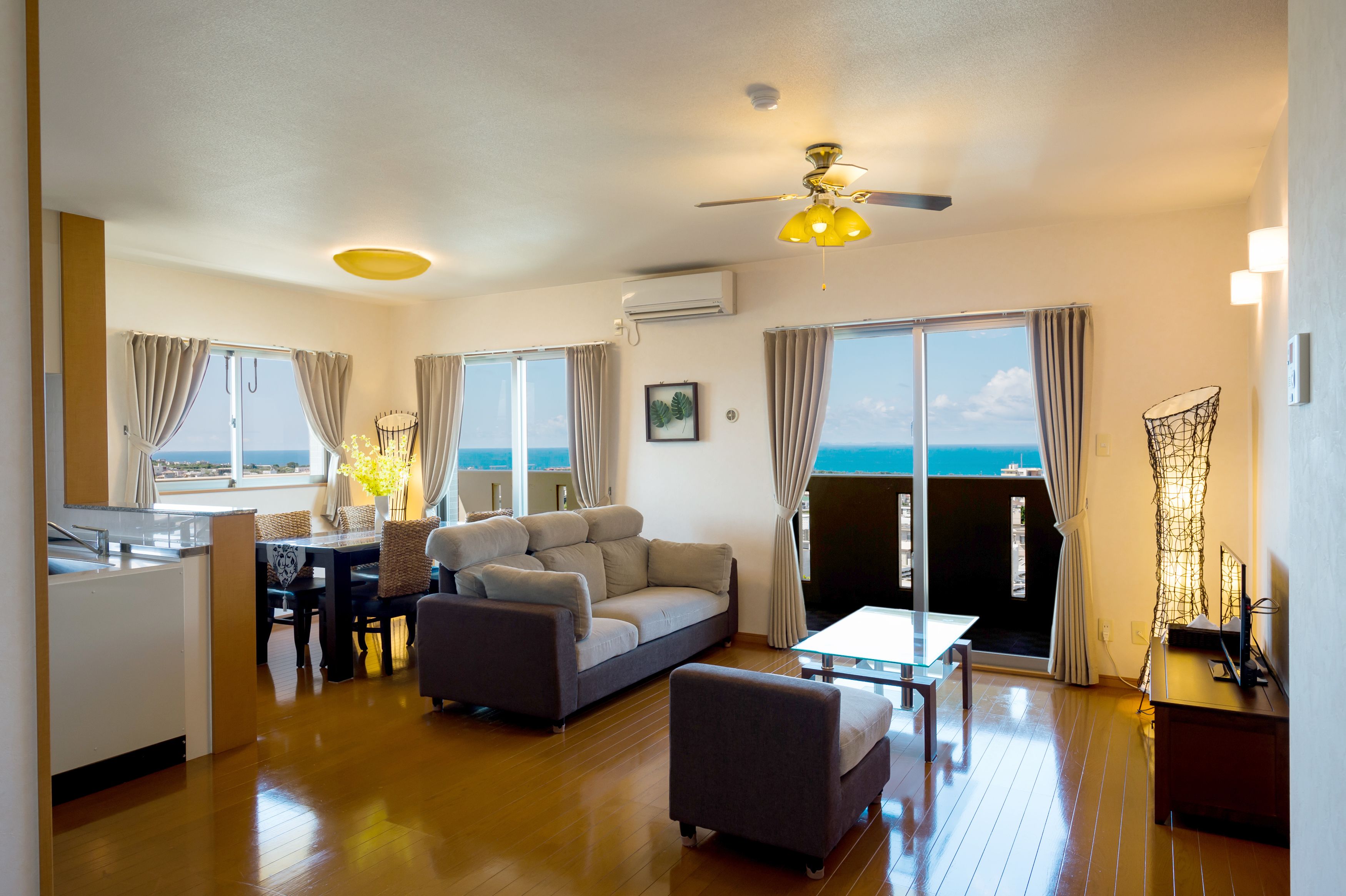 A modern living room with a sofa, armchair, and dining table. There is a ceiling fan, a view of the ocean through large windows, and a kitchen area with a sink and counter.