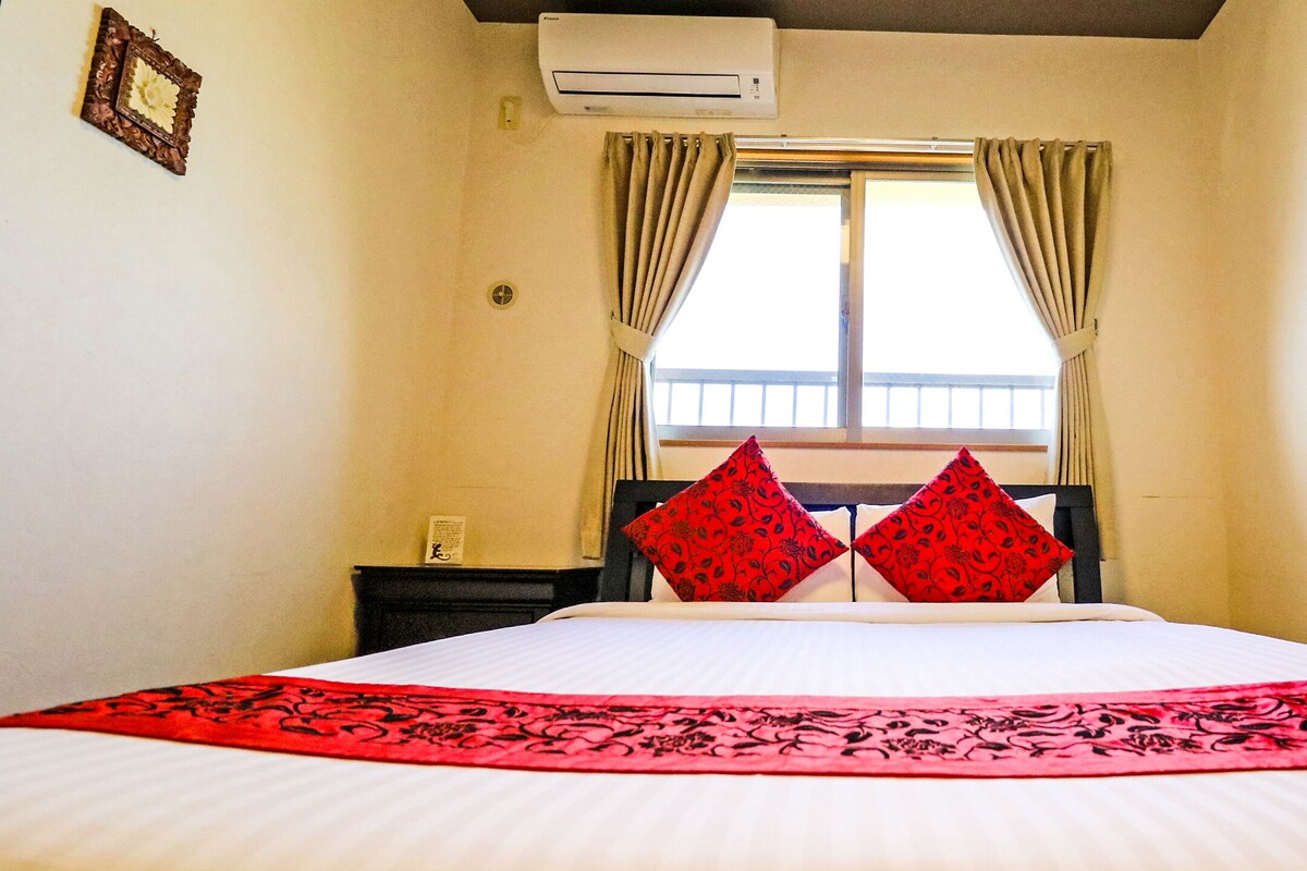 A hotel room with a bed, a window with curtains, an air conditioning unit, and a nightstand.