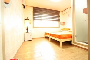 Ein kleiner, heller Raum mit einem Einzelbett, einem Nachttisch, einer Lampe und einem Fenster mit Jalousien.