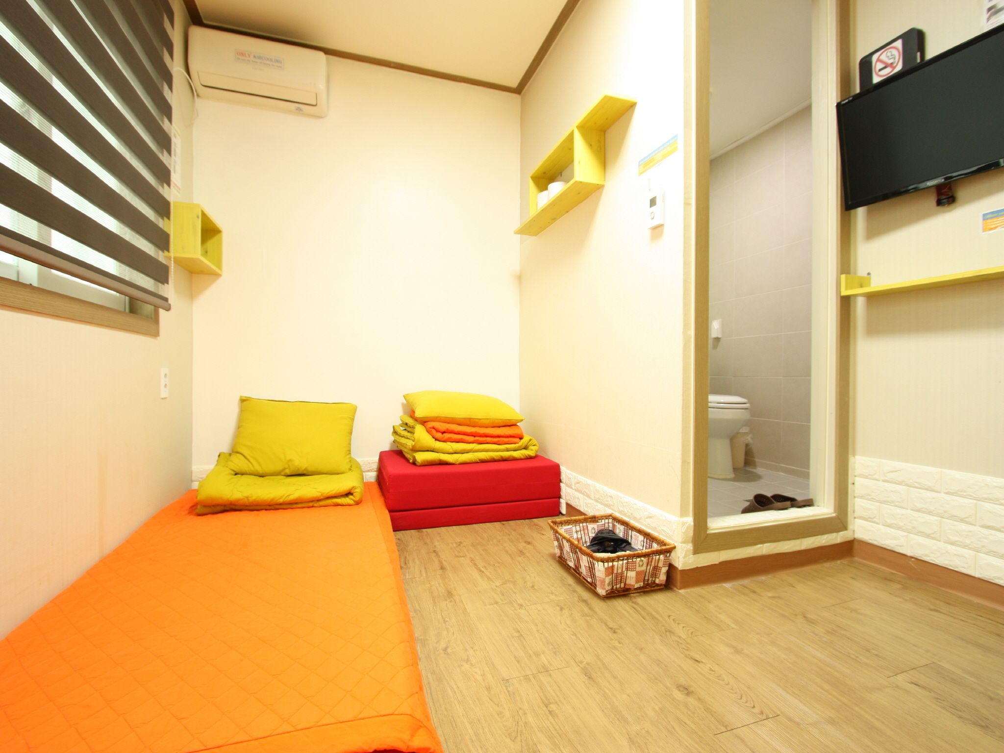 A small, brightly lit room with a single bed, a red bench, and a basket on the floor.