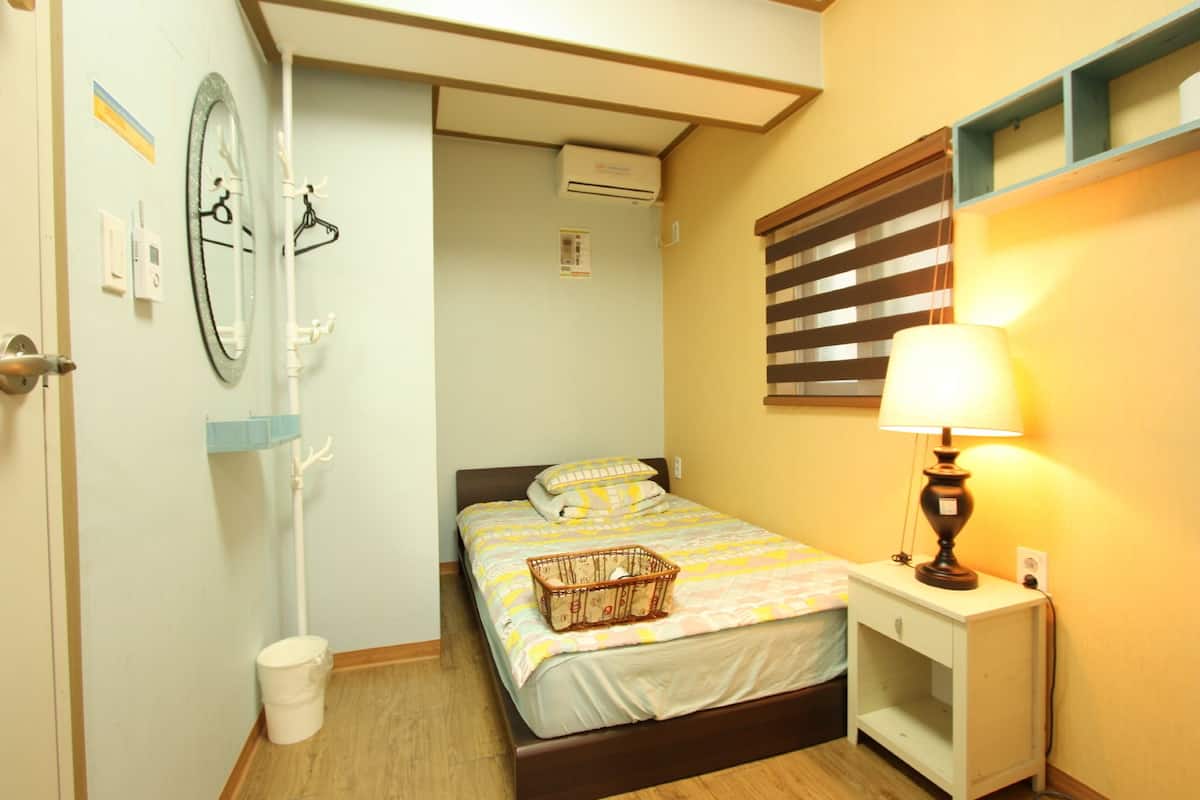 Une petite chambre confortable, avec un lit simple, une table de chevet, une lampe et un climatiseur fixé au mur.