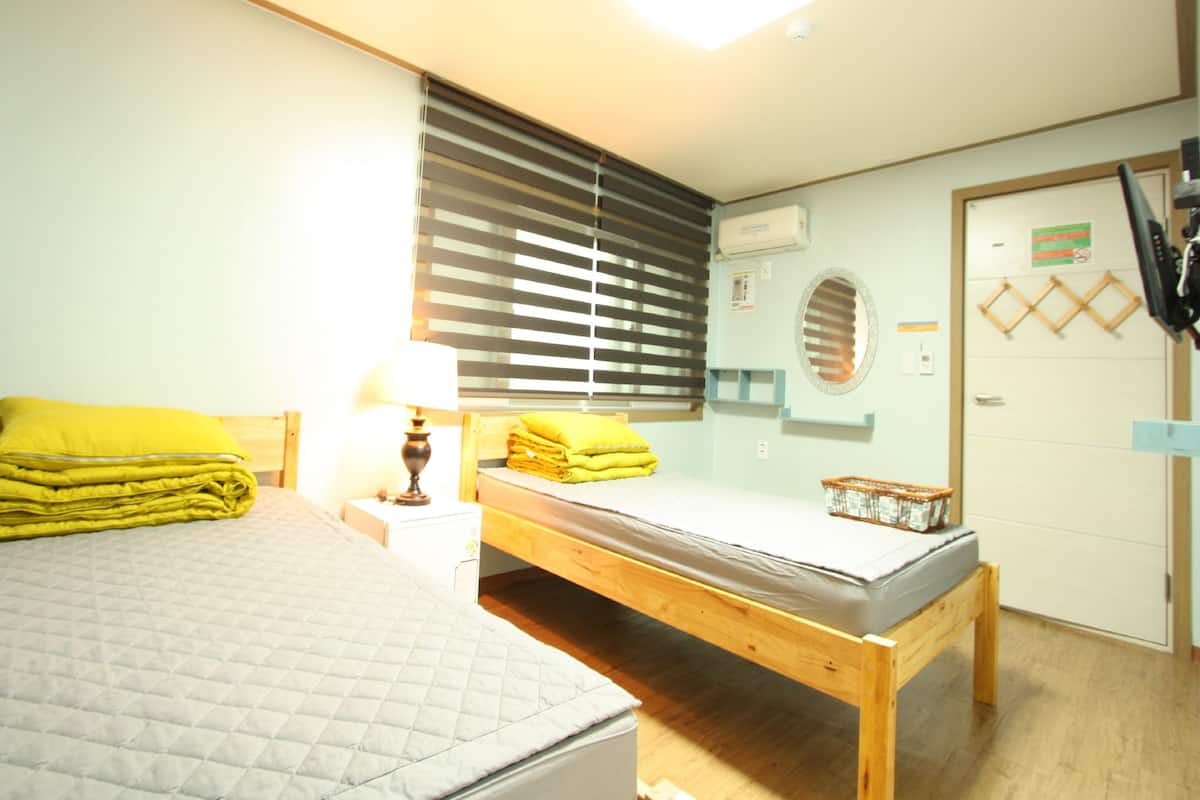 Une chambre à coucher comprenant un lit, une table de chevet avec une lampe, un banc, une fenêtre avec des stores, un appareil de climatisation, un miroir et une porte avec un sac suspendu.