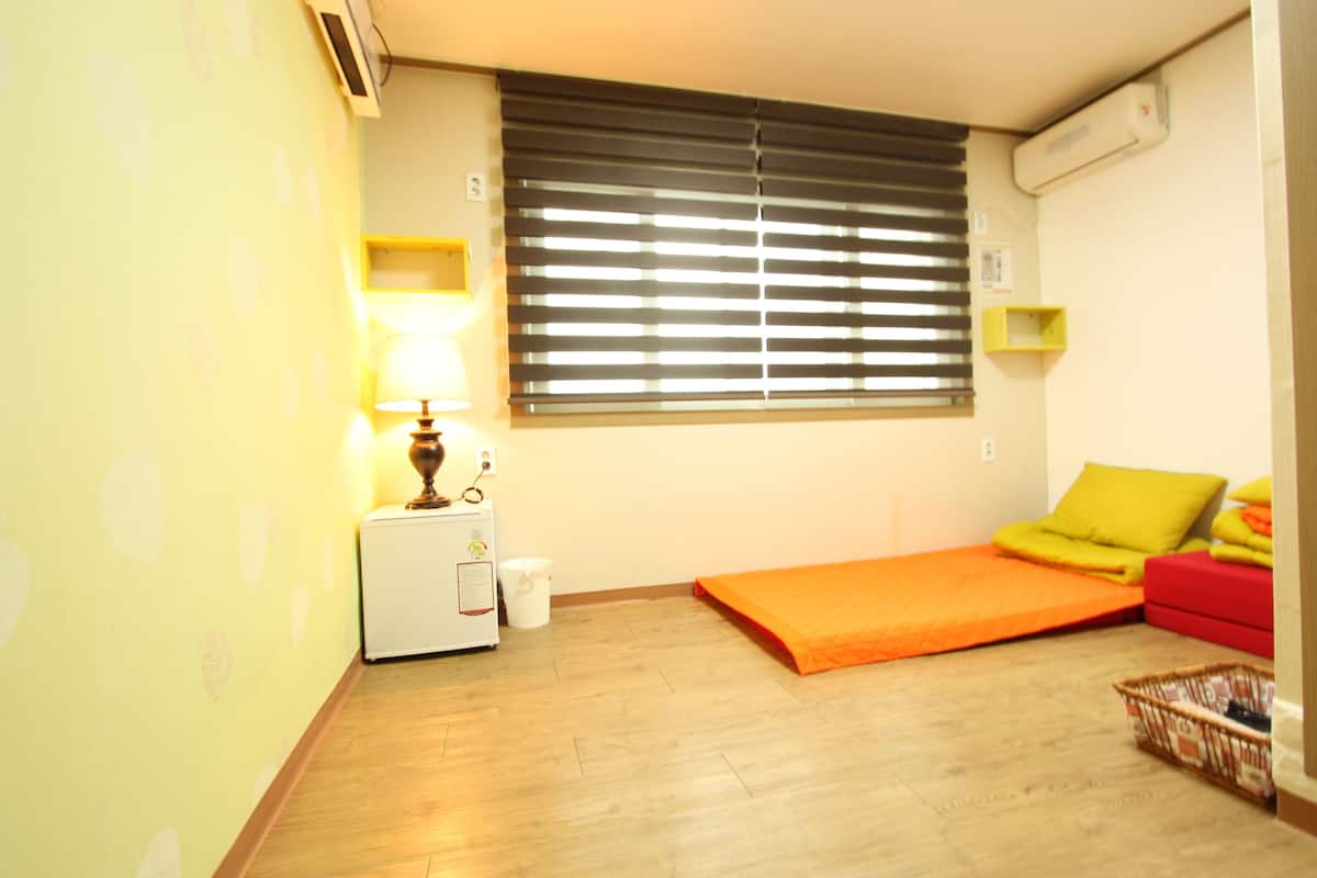 Une chambre avec un lit, une petite table avec une lampe, une fenêtre avec des stores et un climatiseur fixé au mur.