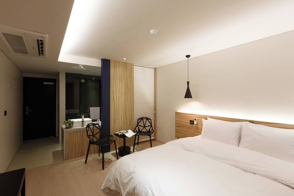 Ein modernes Hotelzimmer mit einem großen Bett, einem Schreibtisch mit Stuhl, einem kleinen Tisch mit Stuhl und einem durchgehenden Türrahmen einsehbaren Badezimmer.