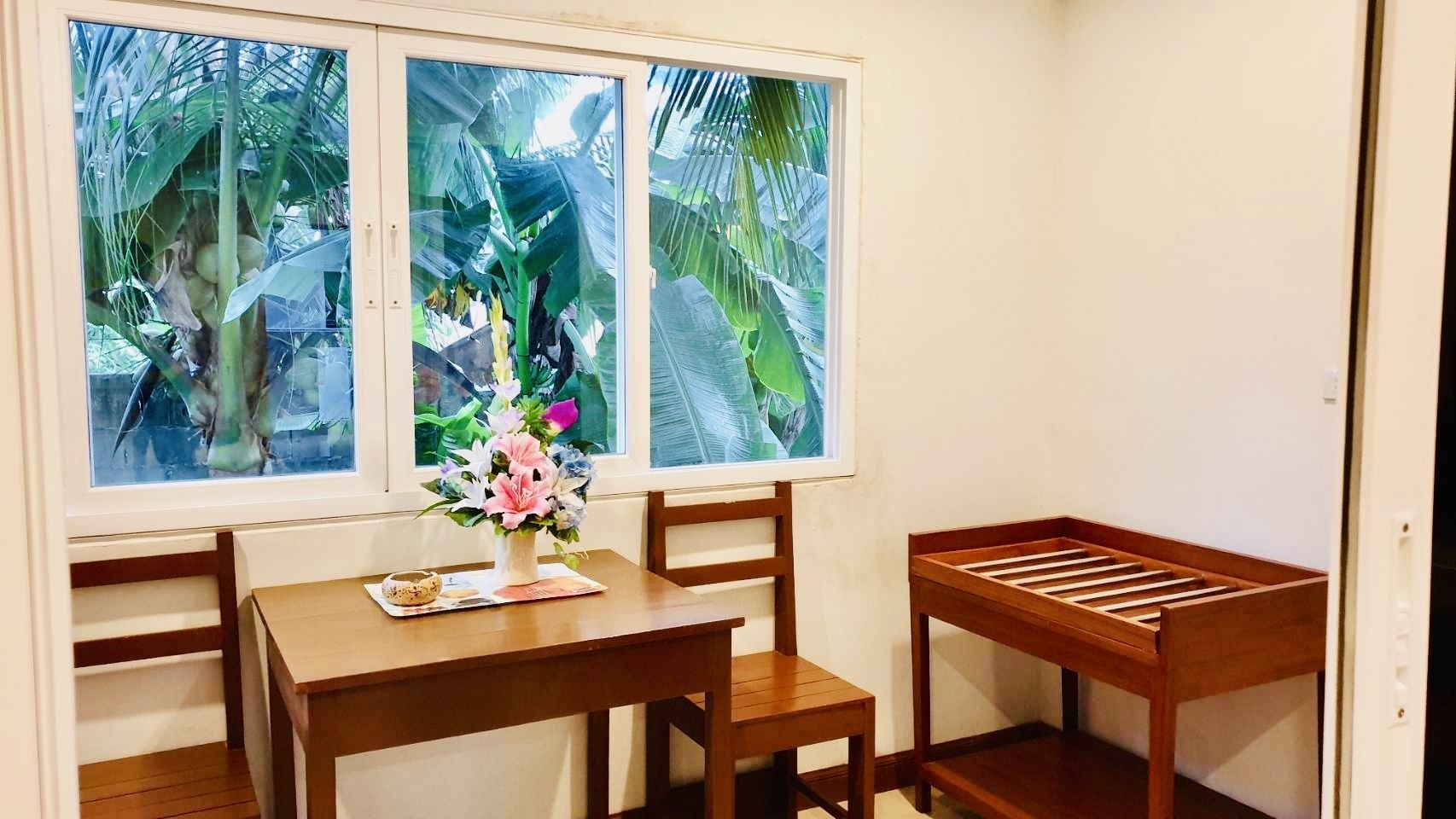 A small room with a wooden table and chairs, a window with a view of tropical plants, and a small wooden bench.