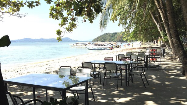 Un restaurante frente a la playa con mesas y sillas, con vista al mar y a las embarcaciones.