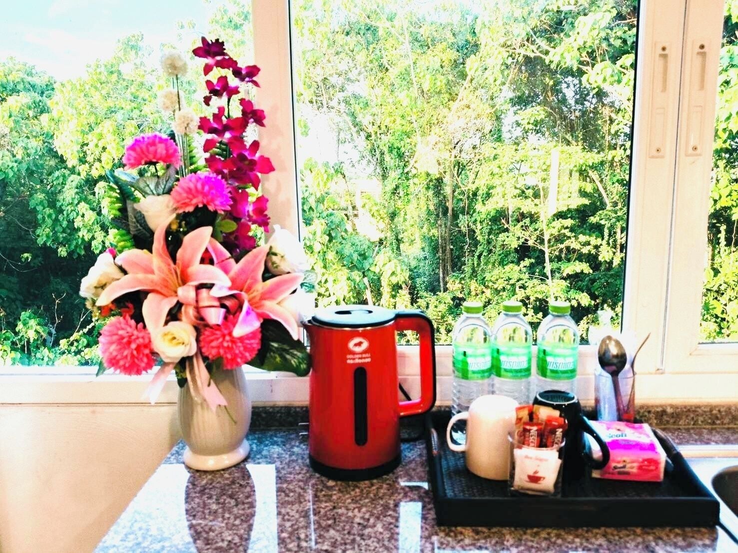 A kitchen counter with a red kettle, a white mug, and a tray with green bottles, a spoon, and packaged snacks.