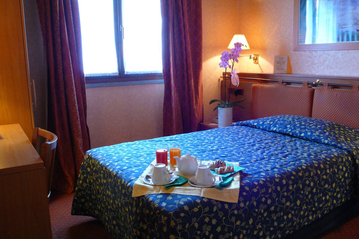 A hotel room with a bed, a table with breakfast items, a lamp, and a window with curtains.