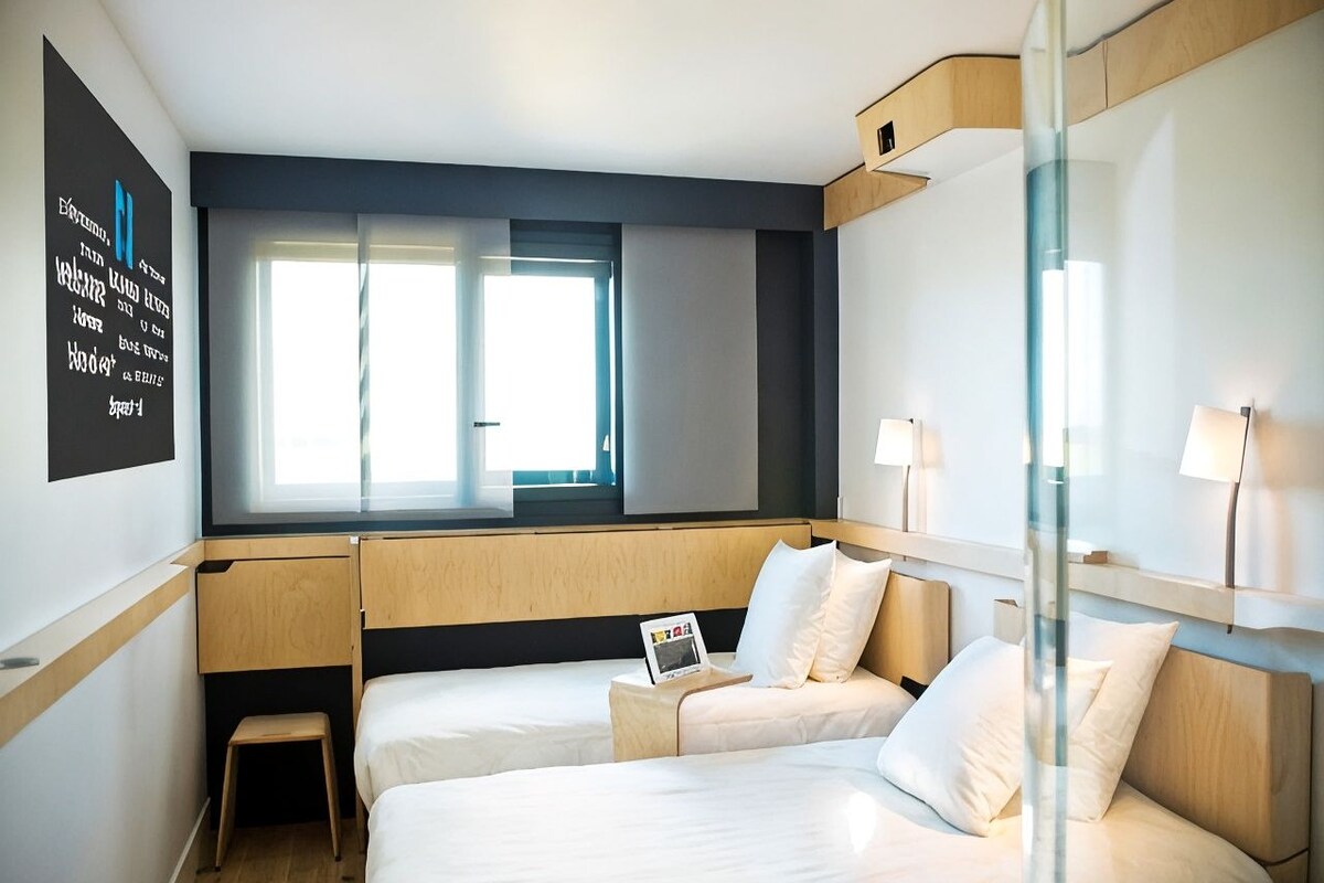 A hotel room with two beds, a small stool, a window, and a wall-mounted TV.