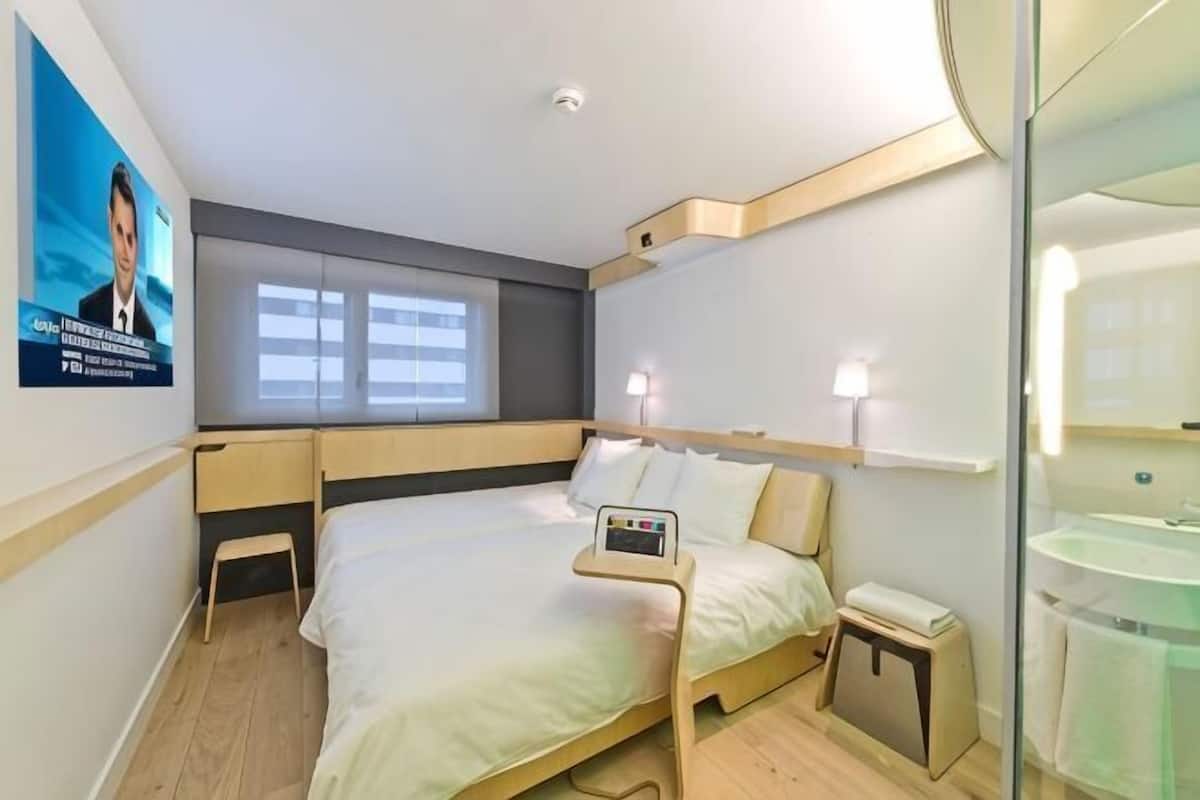 Une chambre d’hôtel comprenant un lit, une table de chevet, une commode et une fenêtre avec des stores.