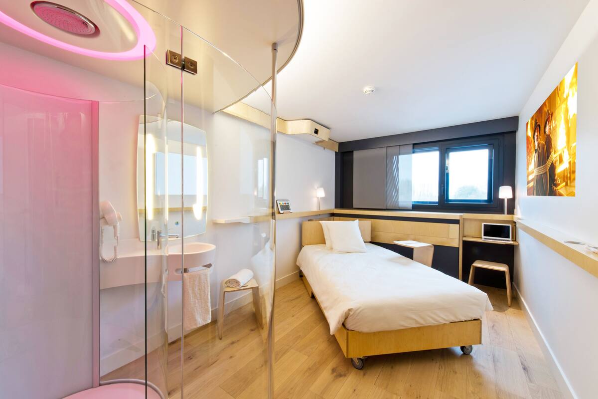 Une chambre d’hôtel moderne dotée d’une douche à parois de verre, d’un lit avec un cadre en bois, d’une table de chevet, d’un bureau et d’une grande fenêtre laissant entrer la lumière naturelle.