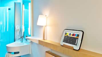 Une chambre d’hôtel comprenant un lit, une table de chevet, une commode, un panneau de commande numérique fixé au mur, une lampe de chevet et une salle de bain avec un lavabo et un miroir.