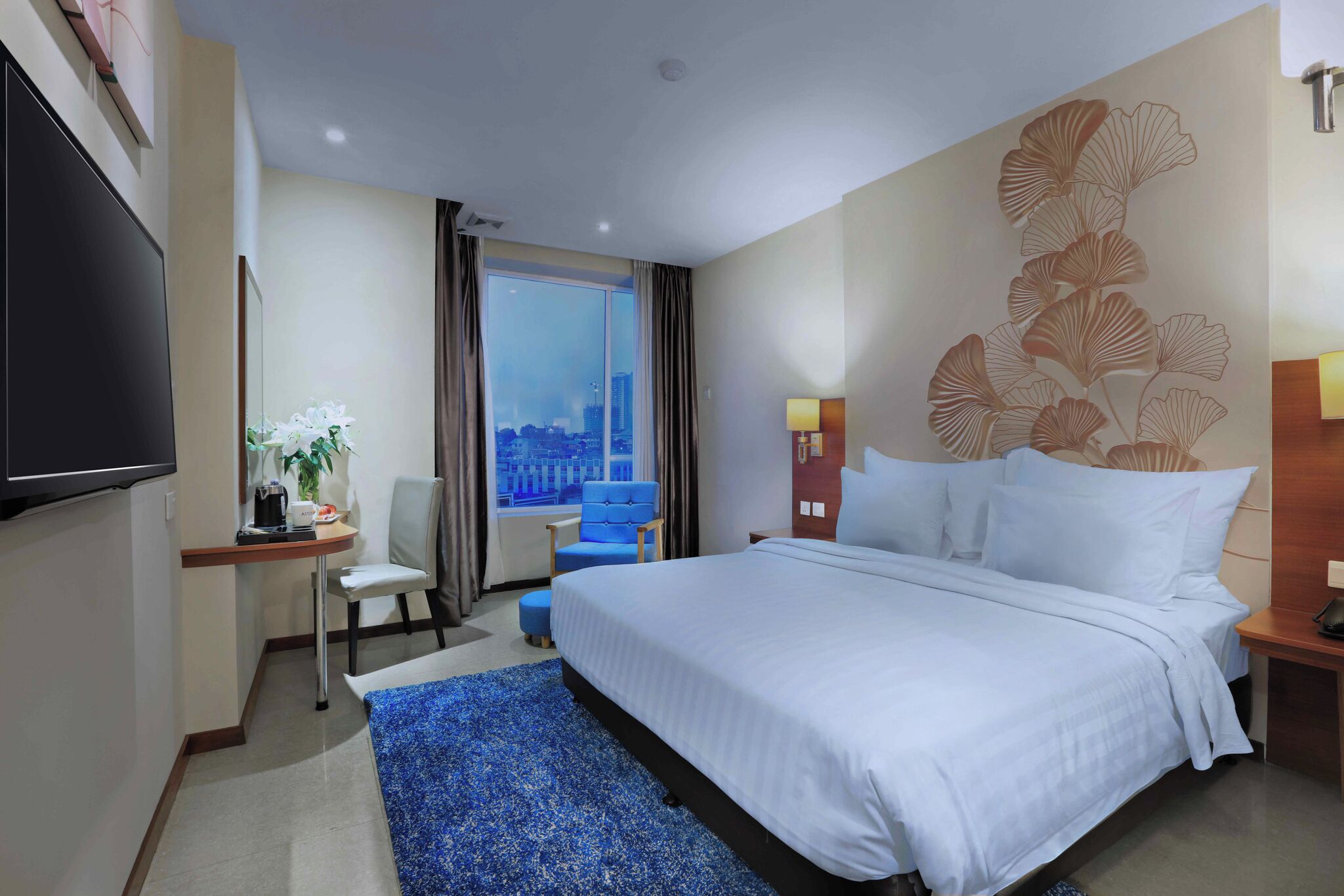 A hotel room with a bed, a desk with a chair, a television, a window with a city view, and a decorative wall.