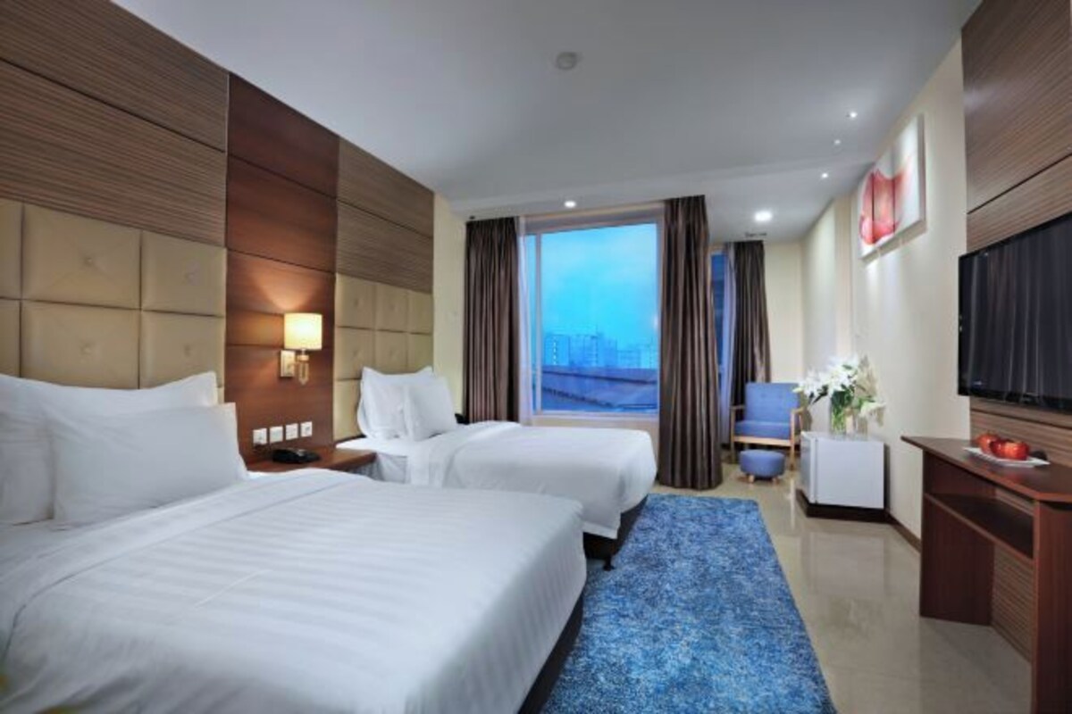 A hotel room with two beds, a large window with a city view, a flat-screen TV, and a desk with a vase of flowers.