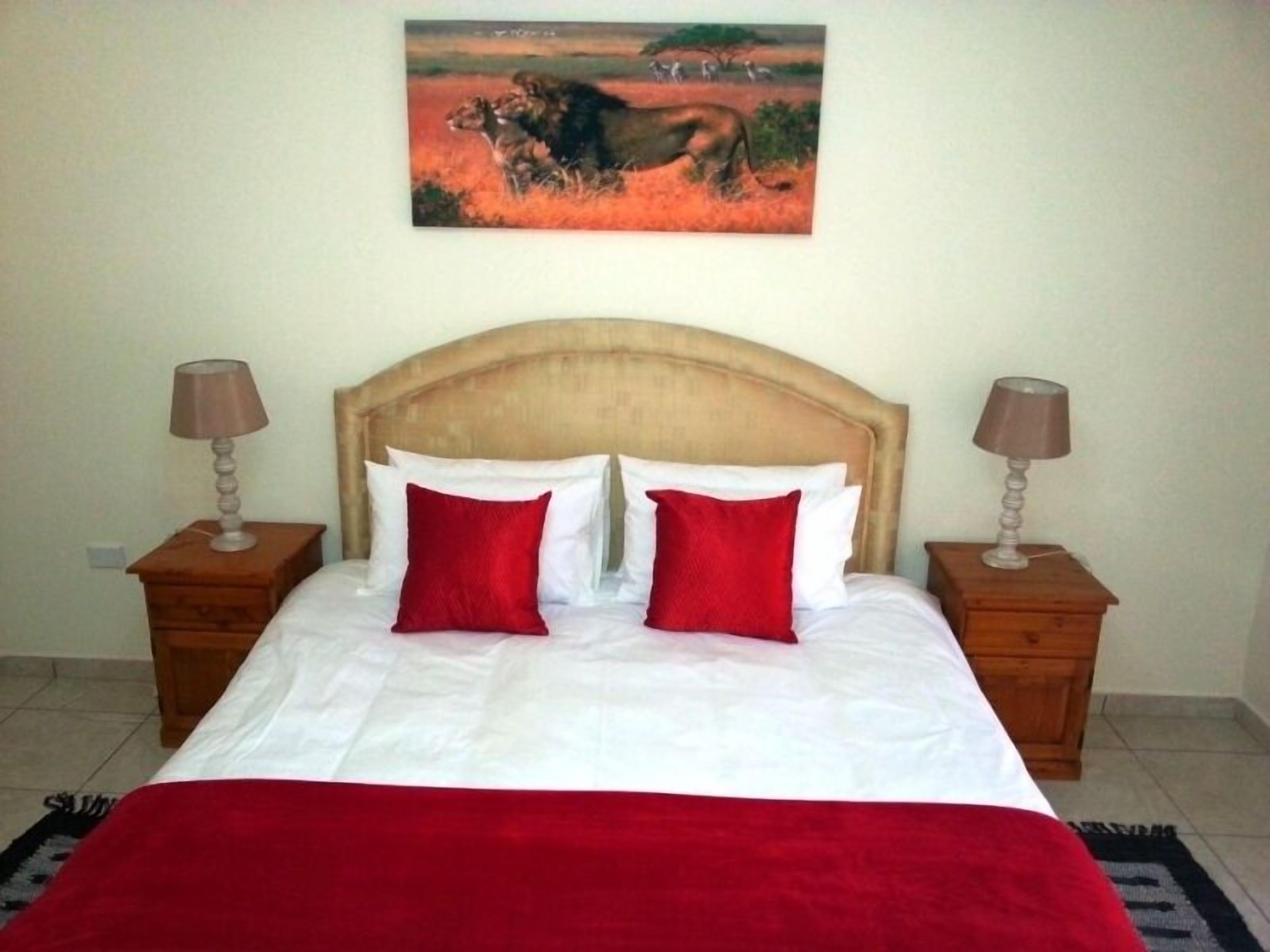 A bed with white bedding and two red pillows, flanked by wooden nightstands with lamps, in a room with a painting of a savannah scene on the wall.