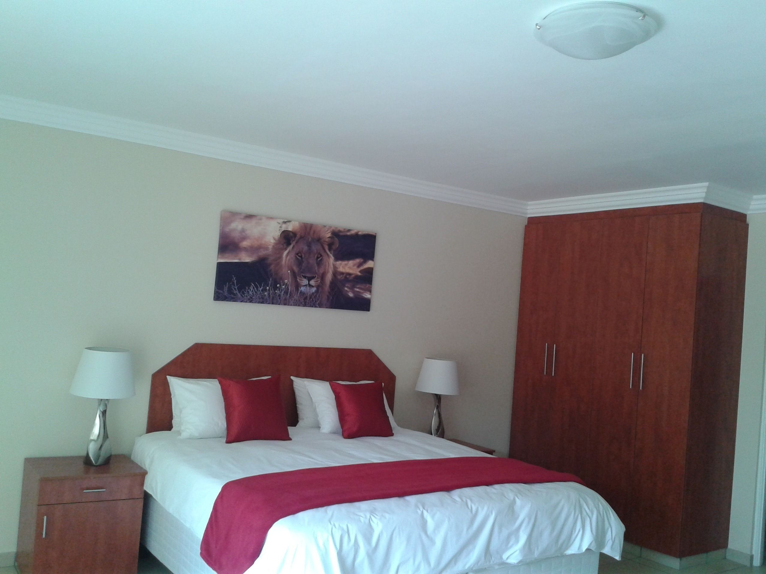 Una habitación de hotel con una cama, dos lámparas de noche, una mesita de noche, un plaid rojo, un armario de madera y un cuadro colgado en la pared con un león.