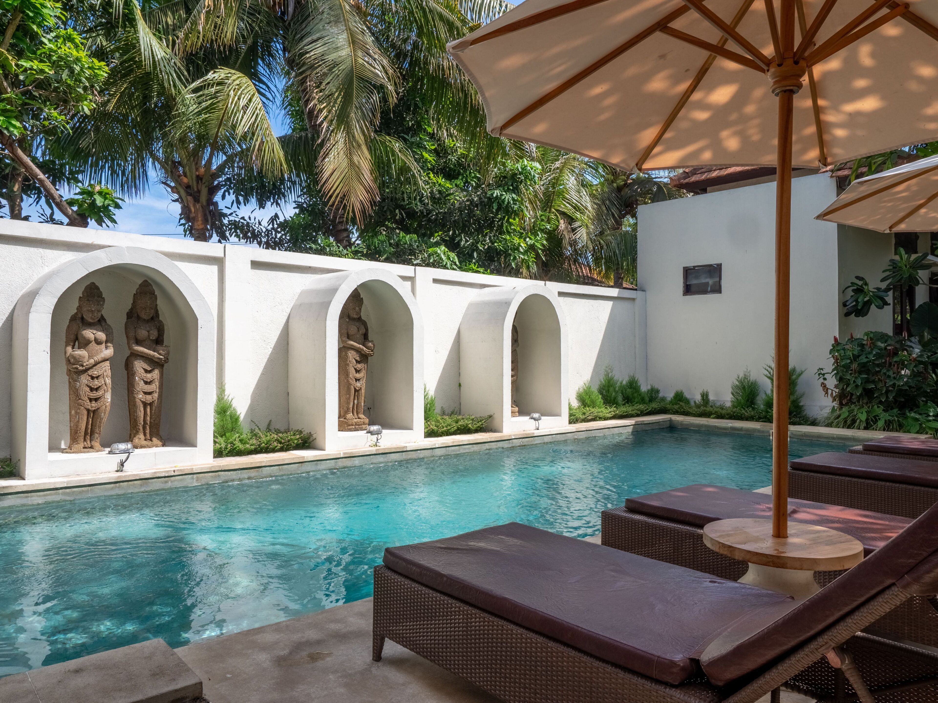 A pool area with lounge chairs and umbrellas, surrounded by white walls and palm trees.