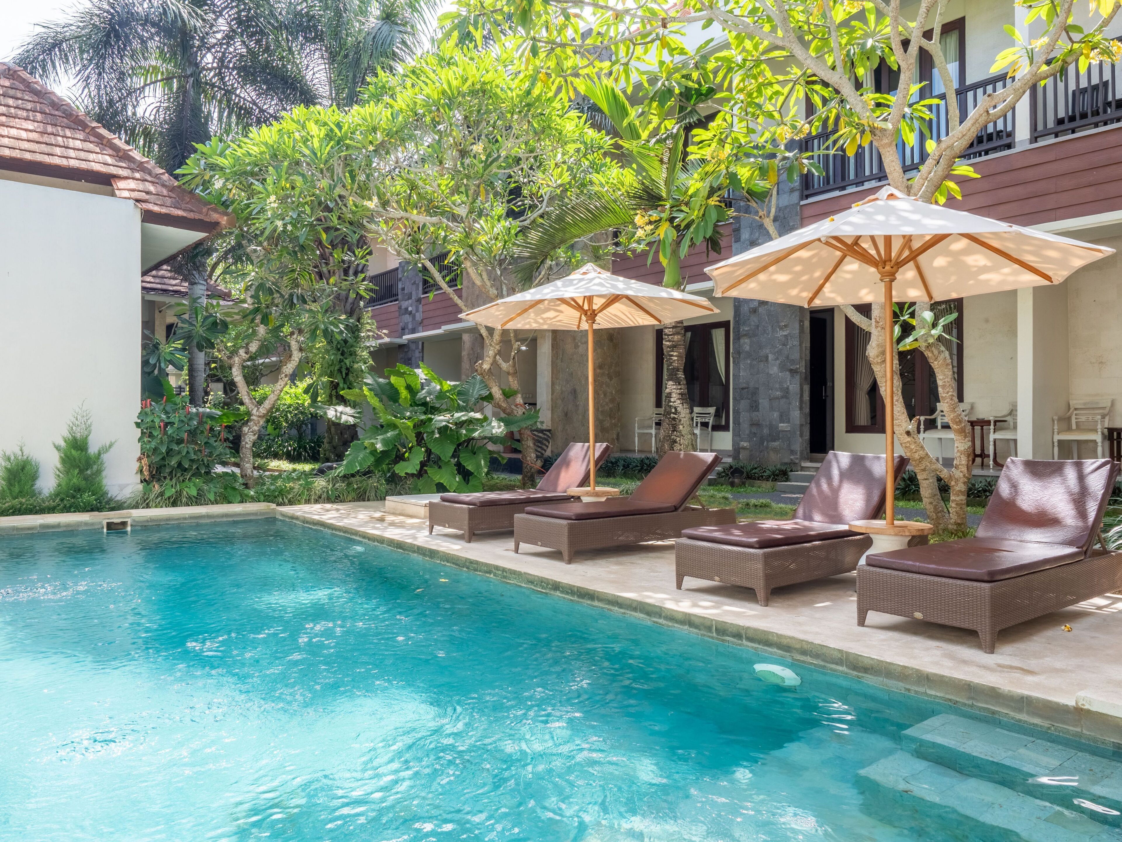 Zona de piscina con sillones de descanso y sombrillas, rodeada de abundante vegetación y un edificio con balcón.