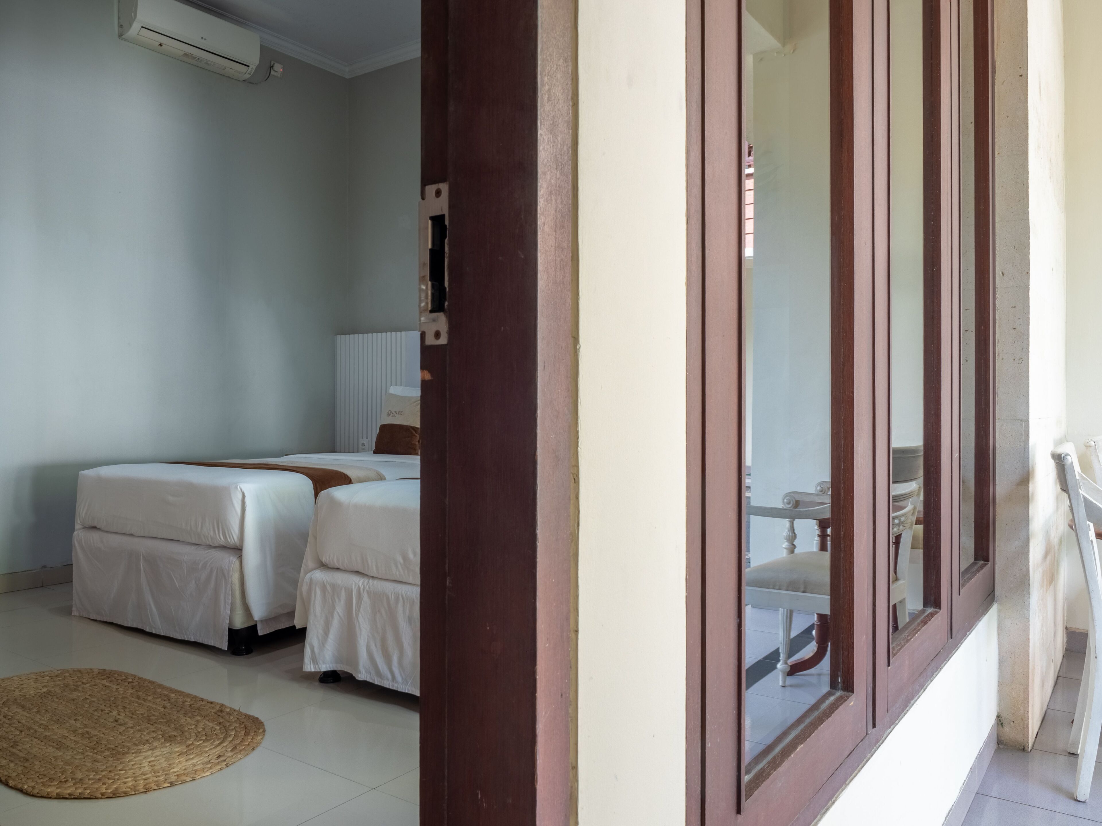Habitación de hotel con dos camas, ventana de madera, unidad de aire acondicionado blanca y un tapete tejido en el suelo.