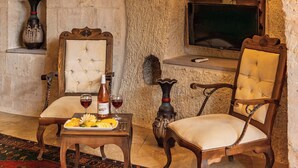 A stone-walled room with a fireplace, a wooden chair, a table with a tray of fruit and wine glasses, and a decorative vase. - Phocas Cave Suites - Special Class (Avanos)