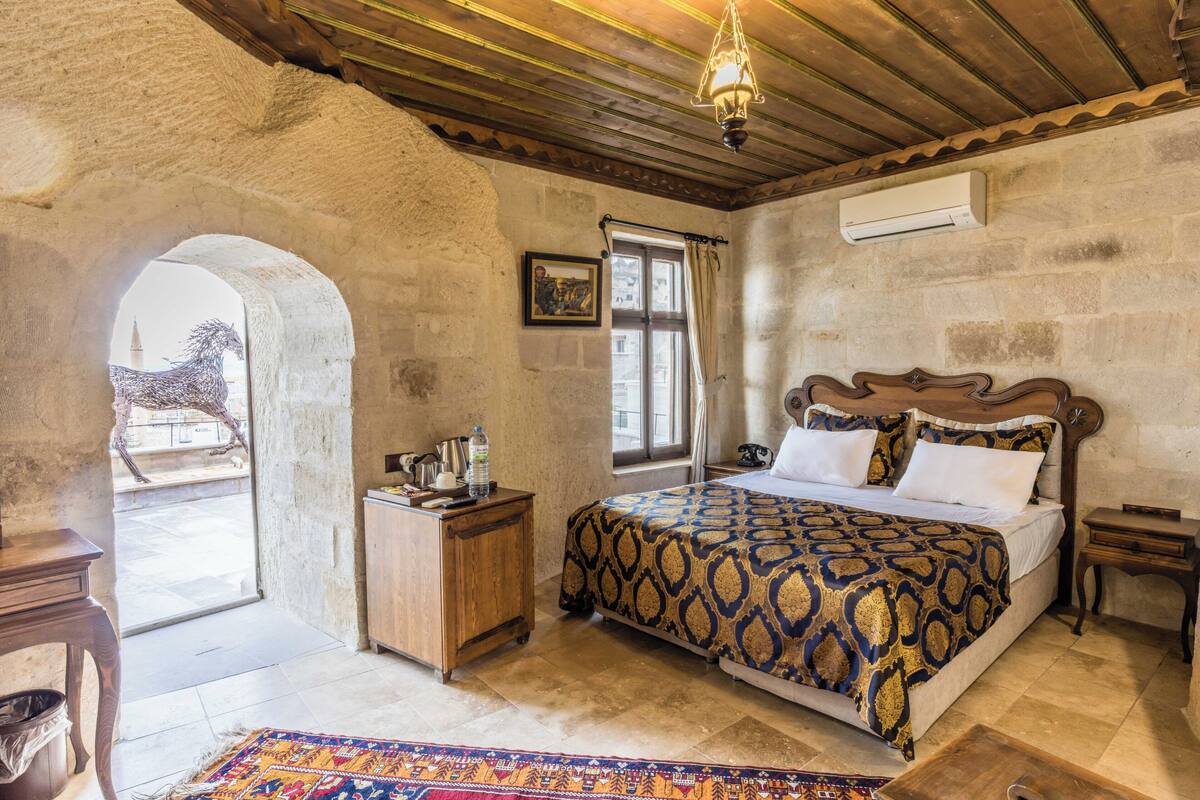 Un dormitorio con pared de piedra, cama de madera, mesita de noche, una pequeña mesa, ventana con cortinas y una lámpara colgante.