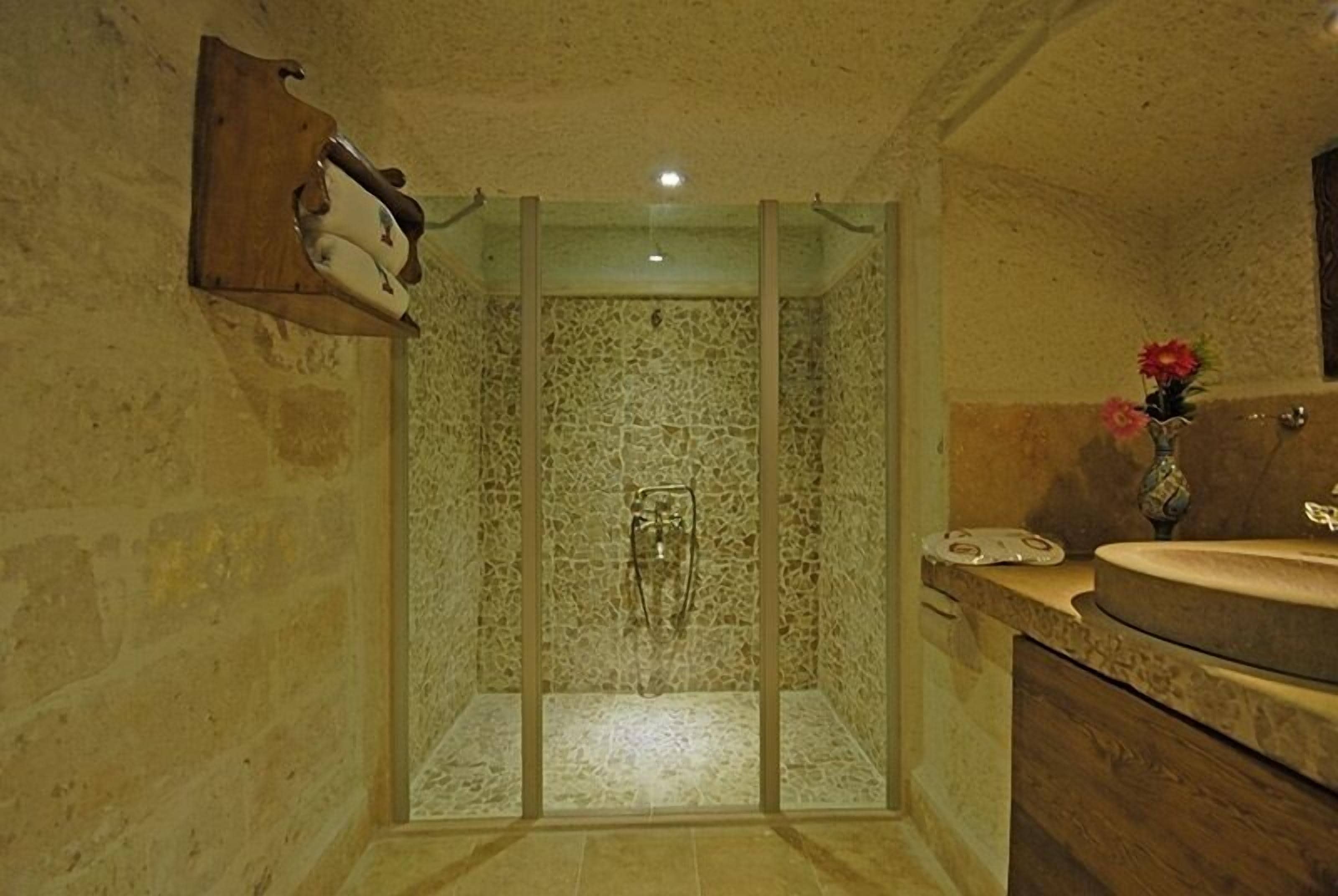 A bathroom with a stone shower enclosure, a wooden shelf with toiletries, and a sink with a vase of flowers.