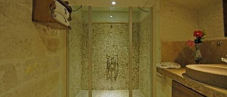 A bathroom with a stone shower enclosure, a wooden shelf with toiletries, and a sink with a vase of flowers.
