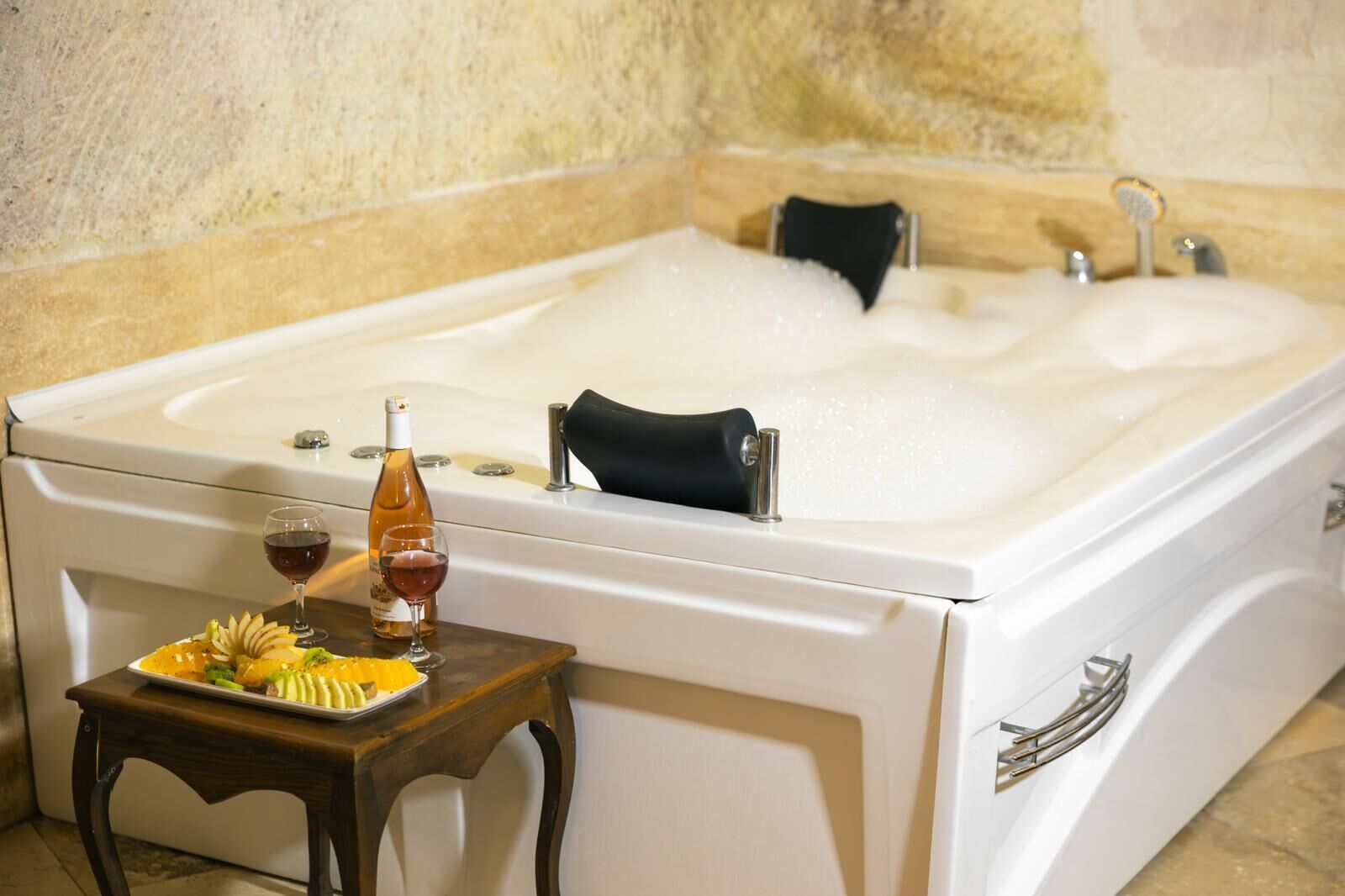 A white bathtub with a black seat, a bottle of wine, and a plate of fruit on a small wooden table beside it.